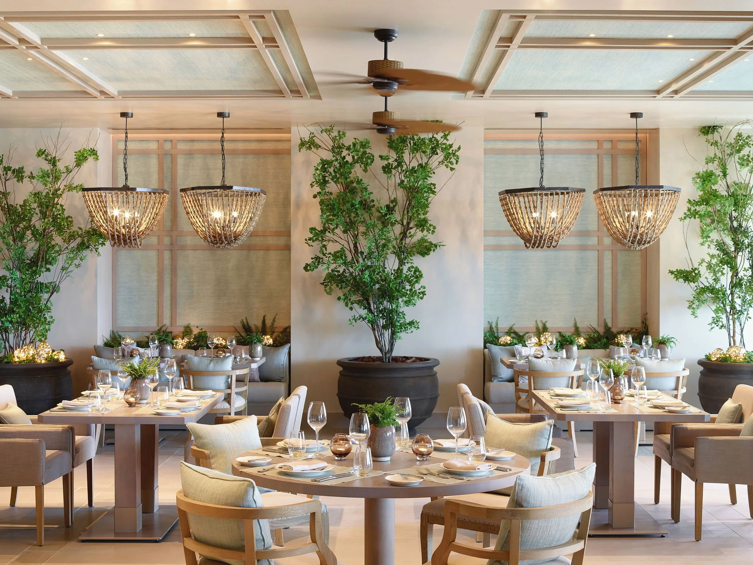 Elegant dining area with round tables decorated with plants and candles, surrounded by light-colored chairs, with large potted plants and hanging chandeliers on the ceiling.
