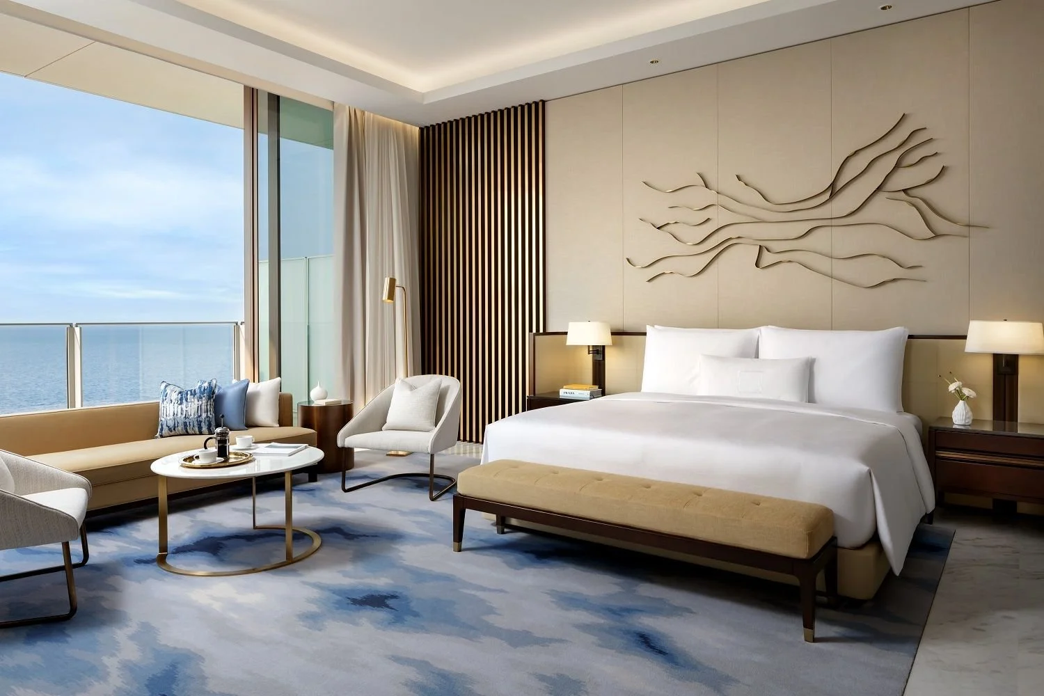 Luxury hotel room with a large bed, bedside tables with lamps, a seating area with a sofa, chairs, and a coffee table by the window overlooking the ocean, decorated with modern art and neutral tones.