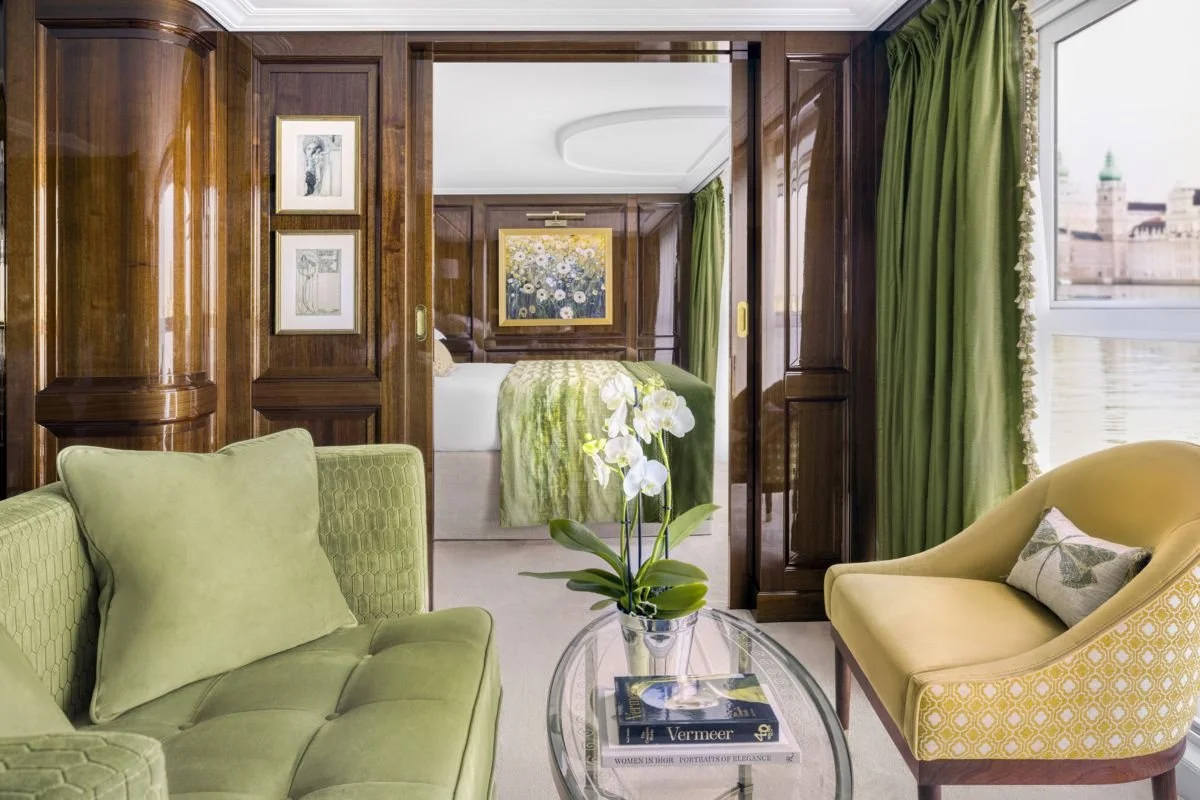 A living area with green and yellow upholstered furniture near a glass table with a potted orchid and books, leading to a bedroom with wooden paneling, framed artwork, and a bed with green bedding and a floral painting above it, with large windows and green curtains.
