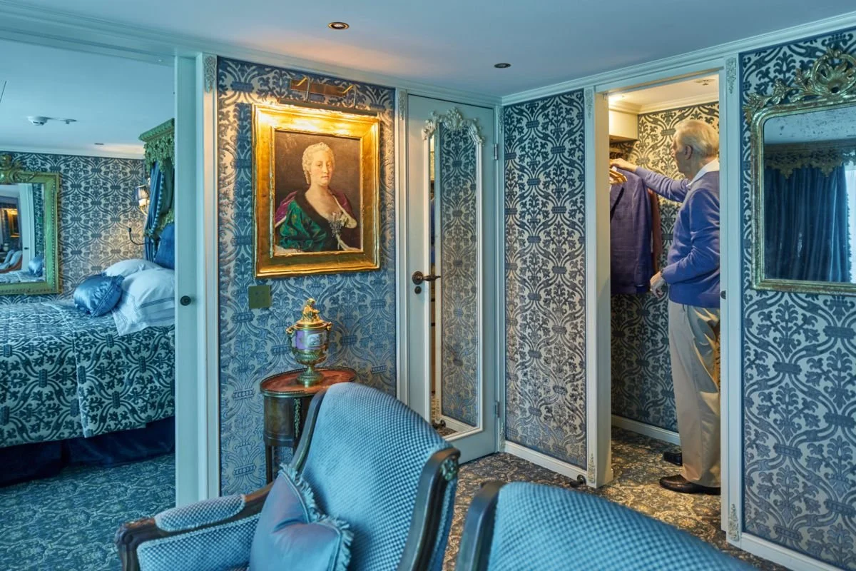 A man is looking at clothes in a walk-in closet inside a luxury bedroom with ornate diamond-patterned wallpaper, gold-framed paintings, and elegant furniture.