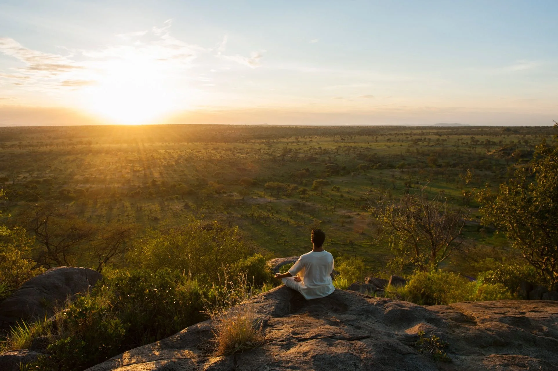 A person practicing yoga or meditation in a seated pose on a rock, overlooking a vast landscape of open fields and scattered trees during sunset or sunrise.