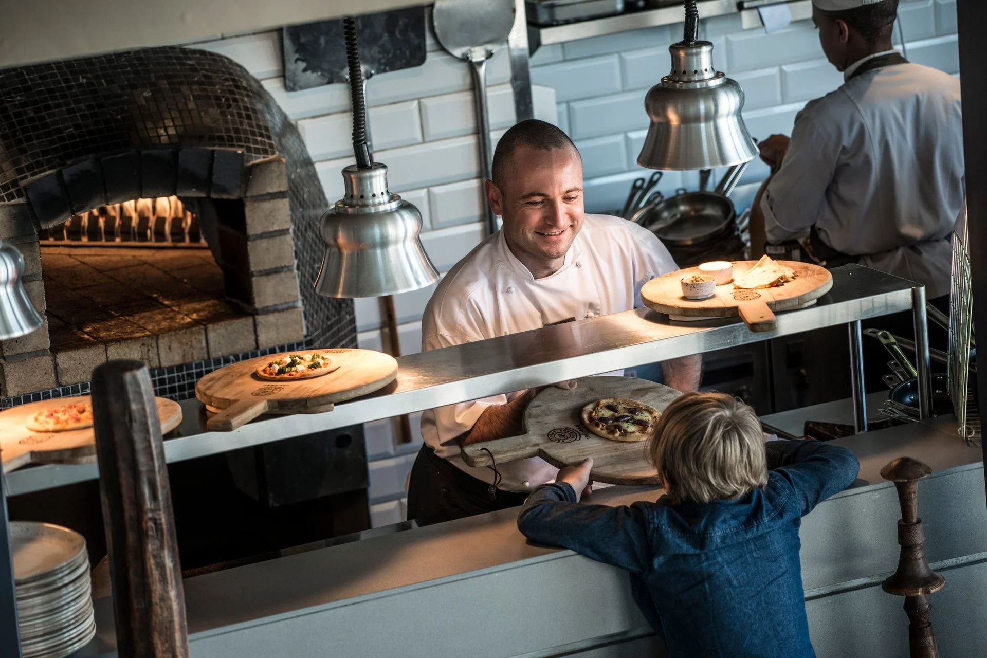 Chef serving pizza to a child behind a kitchen counter in a restaurant with pizza oven in the background.