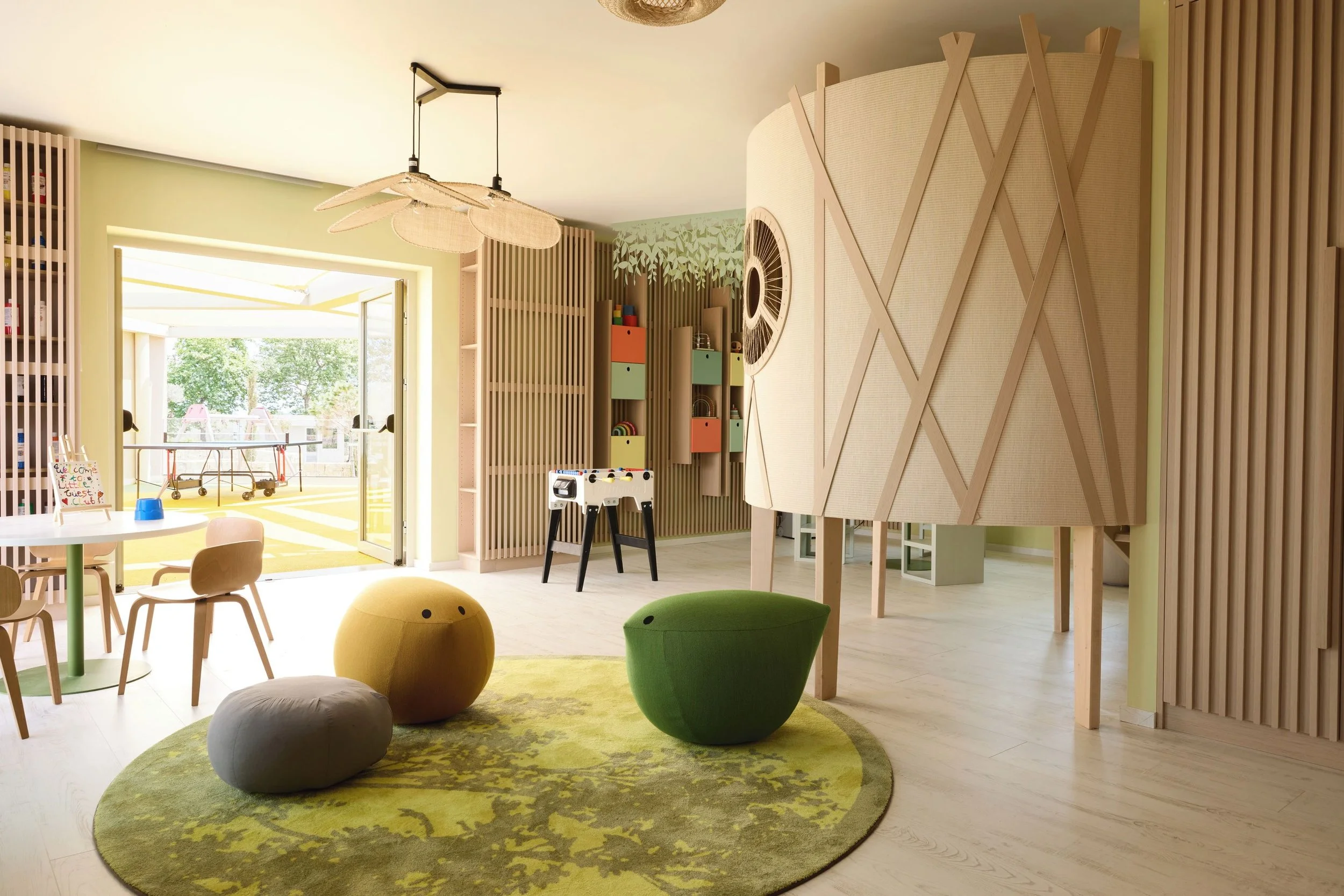 Brightly lit children's playroom with a large oval green rug, bean bag chairs, a small table with chairs, and a sliding glass door leading to an outdoor balcony with trees. The room features wooden slats and playful furniture, with a large, curved, textured wooden structure on the right side.