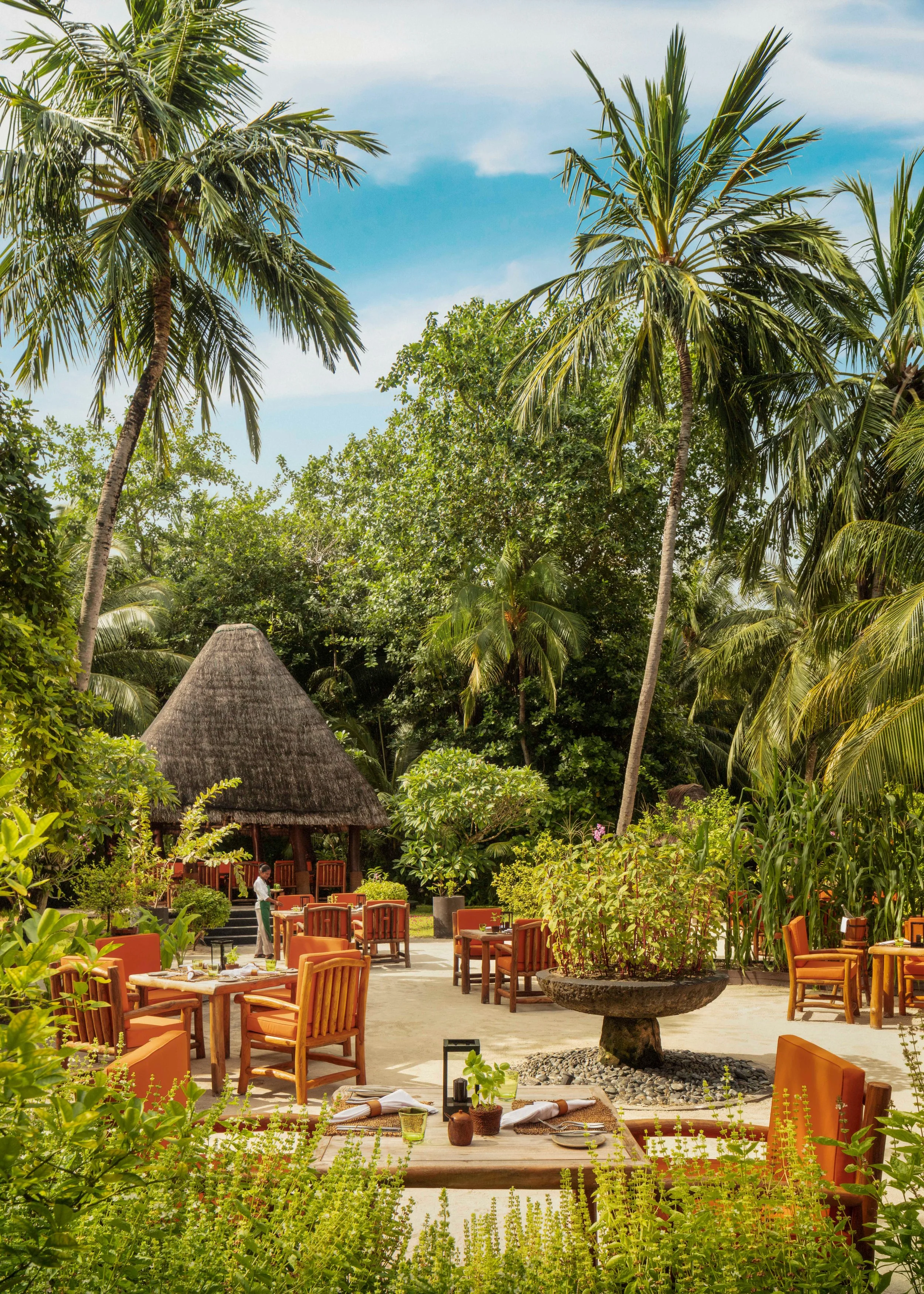 Outdoor restaurant with wooden tables and orange chairs, surrounded by lush green trees and plants, with a thatched roof hut in the background and a fountain at the center, under a partly cloudy sky.