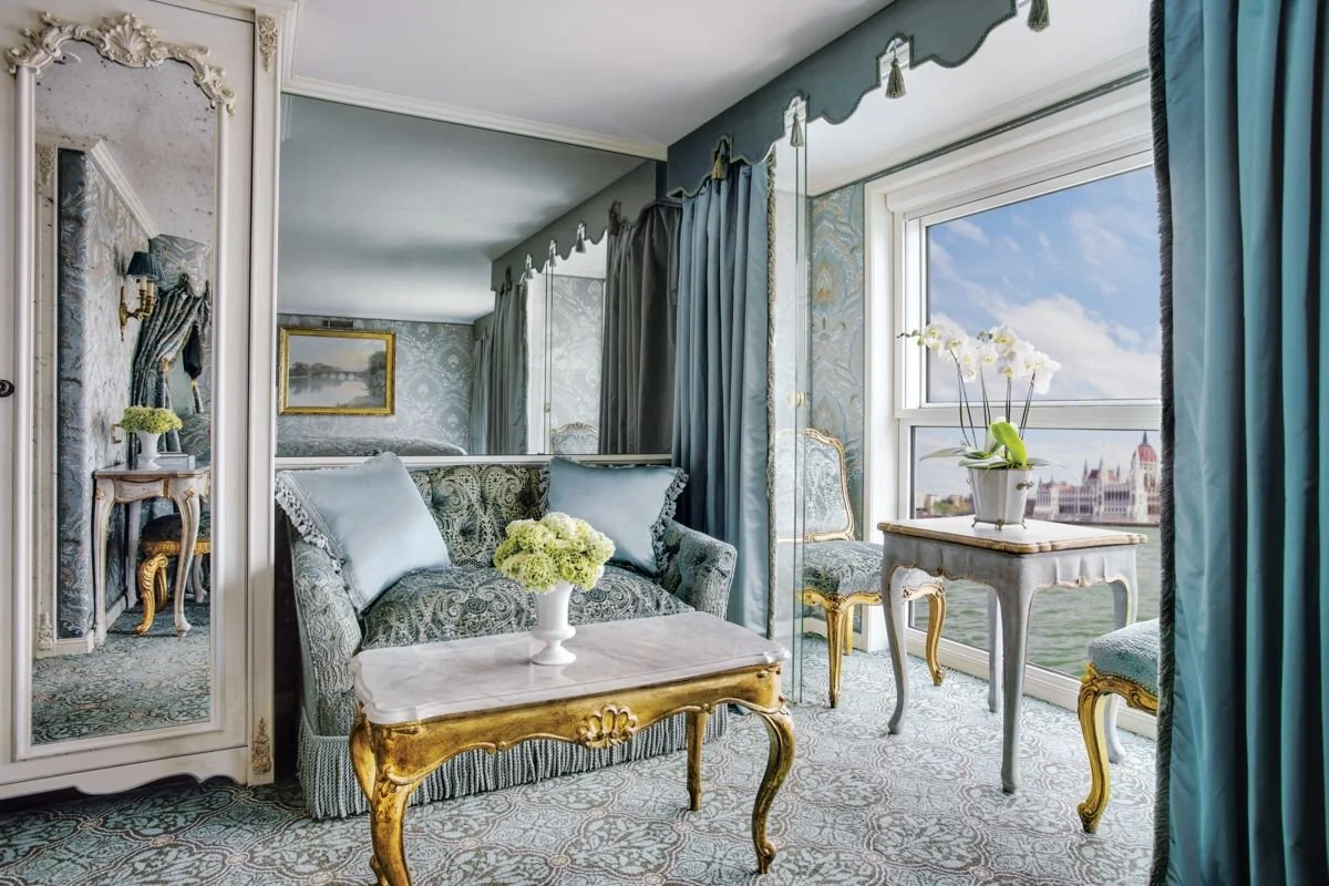 Elegant living room with large window showing city skyline, decorated with ornate vintage furniture, floral arrangements, and blue drapes.