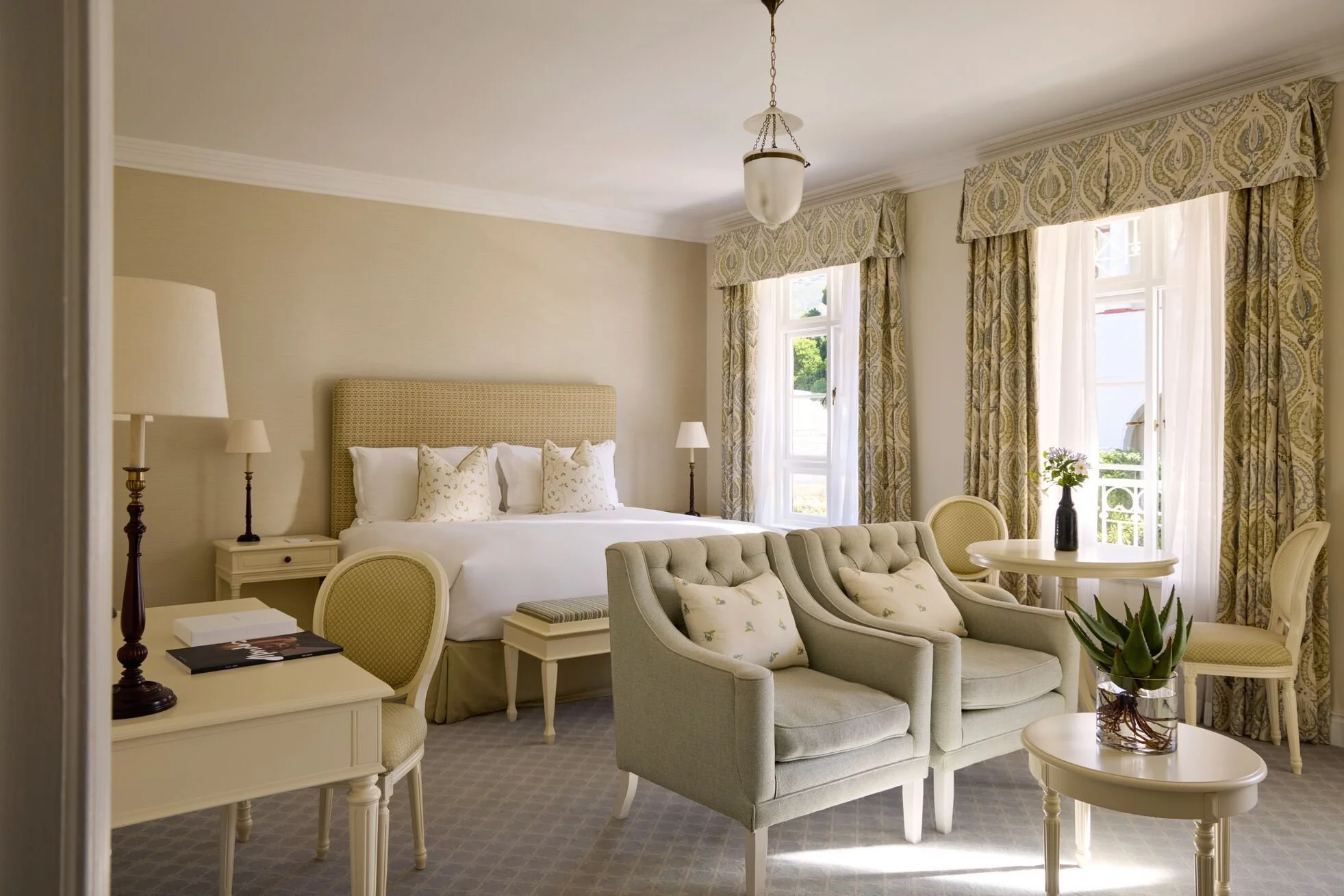A luxurious hotel room with a large bed, beige upholstered headboard, and white linens. There are three patterned pillows on the bed. Beside the bed, there are white nightstands with small lamps. The room has tall windows with patterned curtains, allowing natural light in. In front of the bed, there is a sitting area with two light gray tufted armchairs, a small round table with a plant, and a few decorative pillows. The walls are painted light beige, and the ceiling has crown molding.
