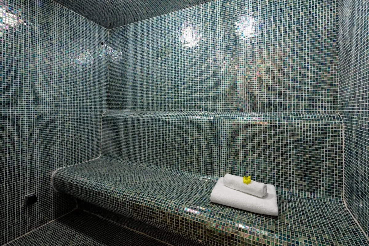 A steam room or sauna interior with walls, ceiling, and bench covered in small iridescent mosaic tiles in various shades of blue and green, with a folded white towel and a small rubber yellow duck placed on top.