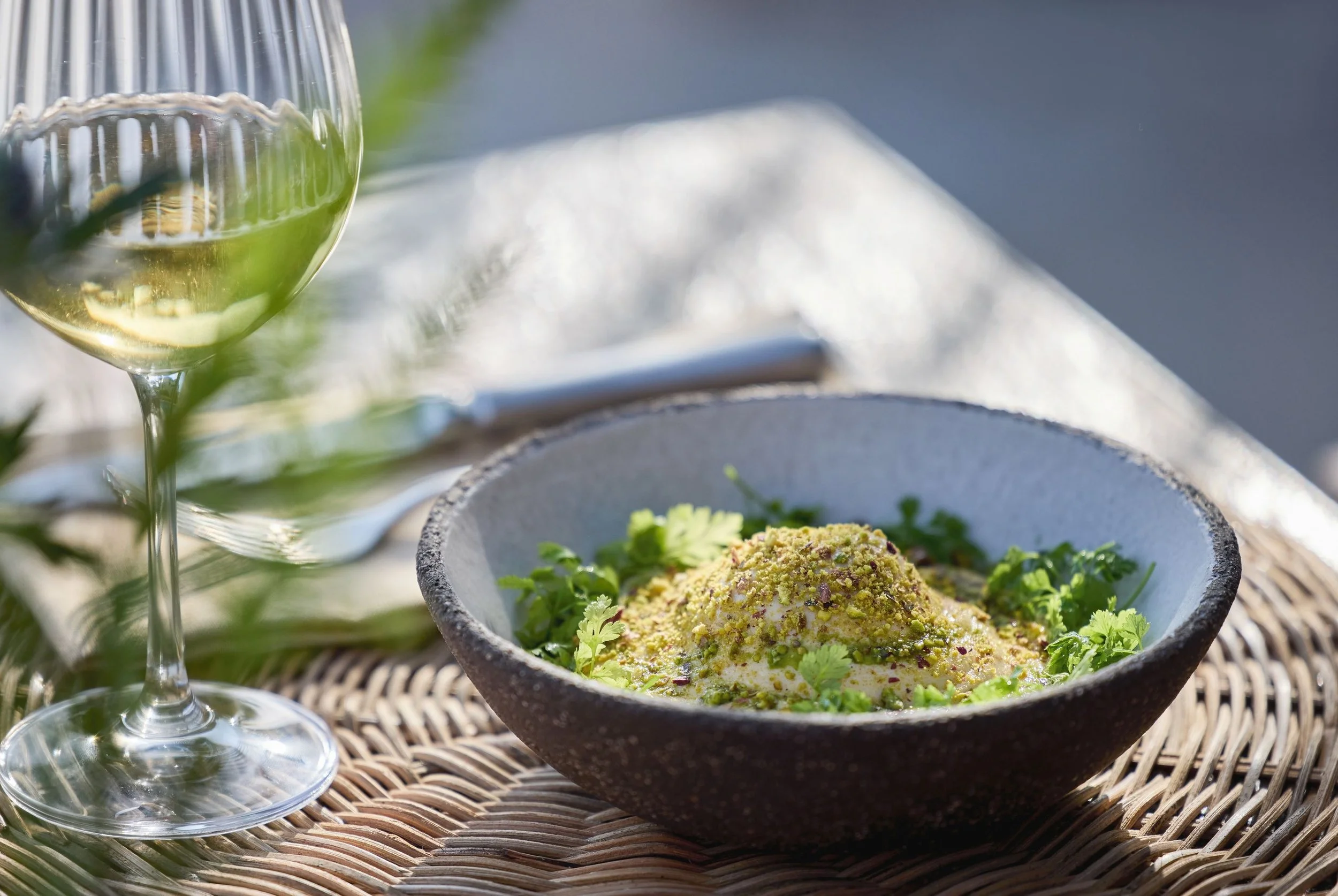 A glass of white wine and a bowl of food garnished with herbs on a woven placemat outdoors.