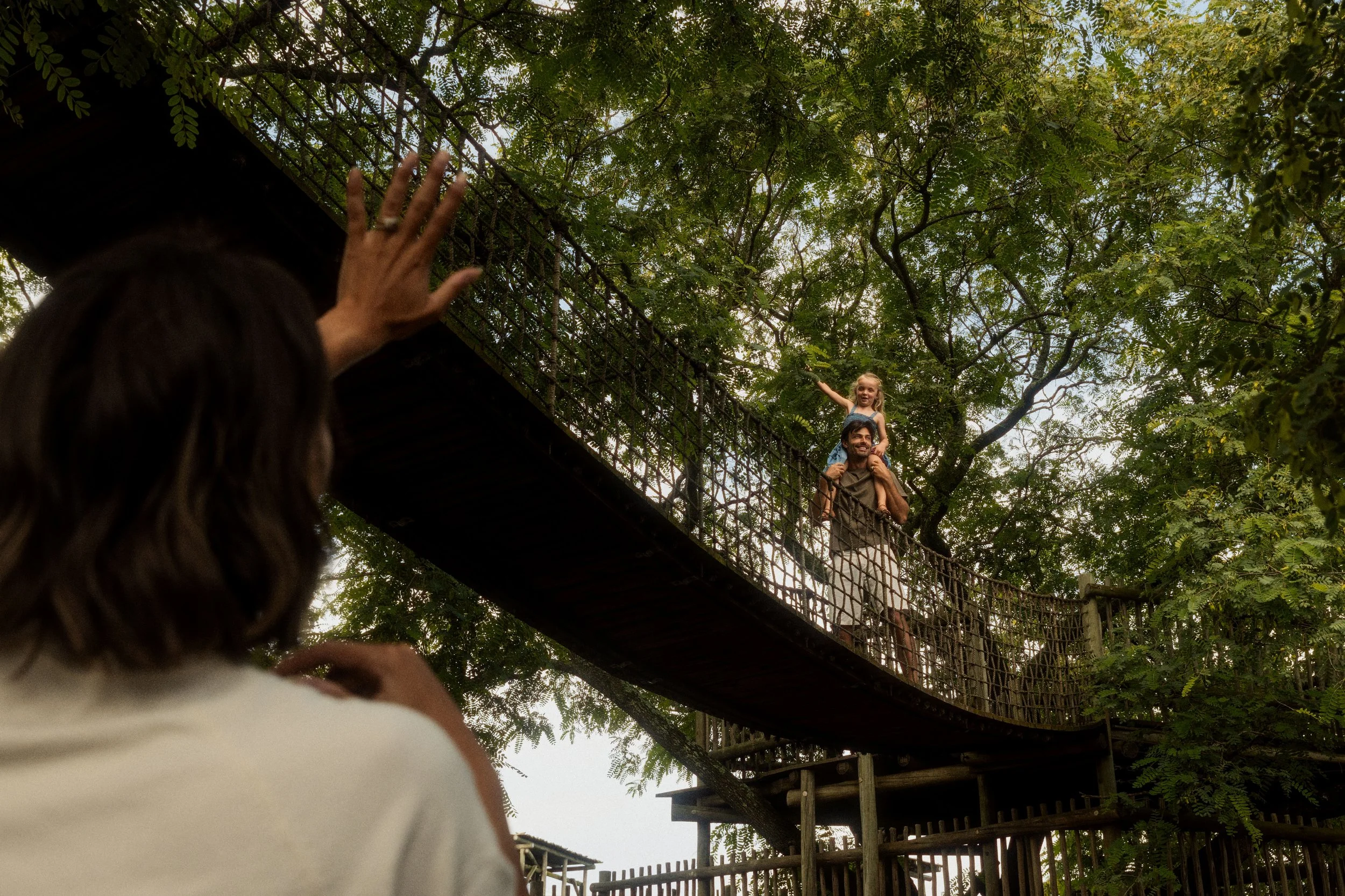 A person with long dark hair is holding a camera up to photograph a man and a young girl on a bridge in a lush, green, outdoor setting. The man is carrying the girl on his shoulders, and both are smiling, surrounded by leafy trees.
