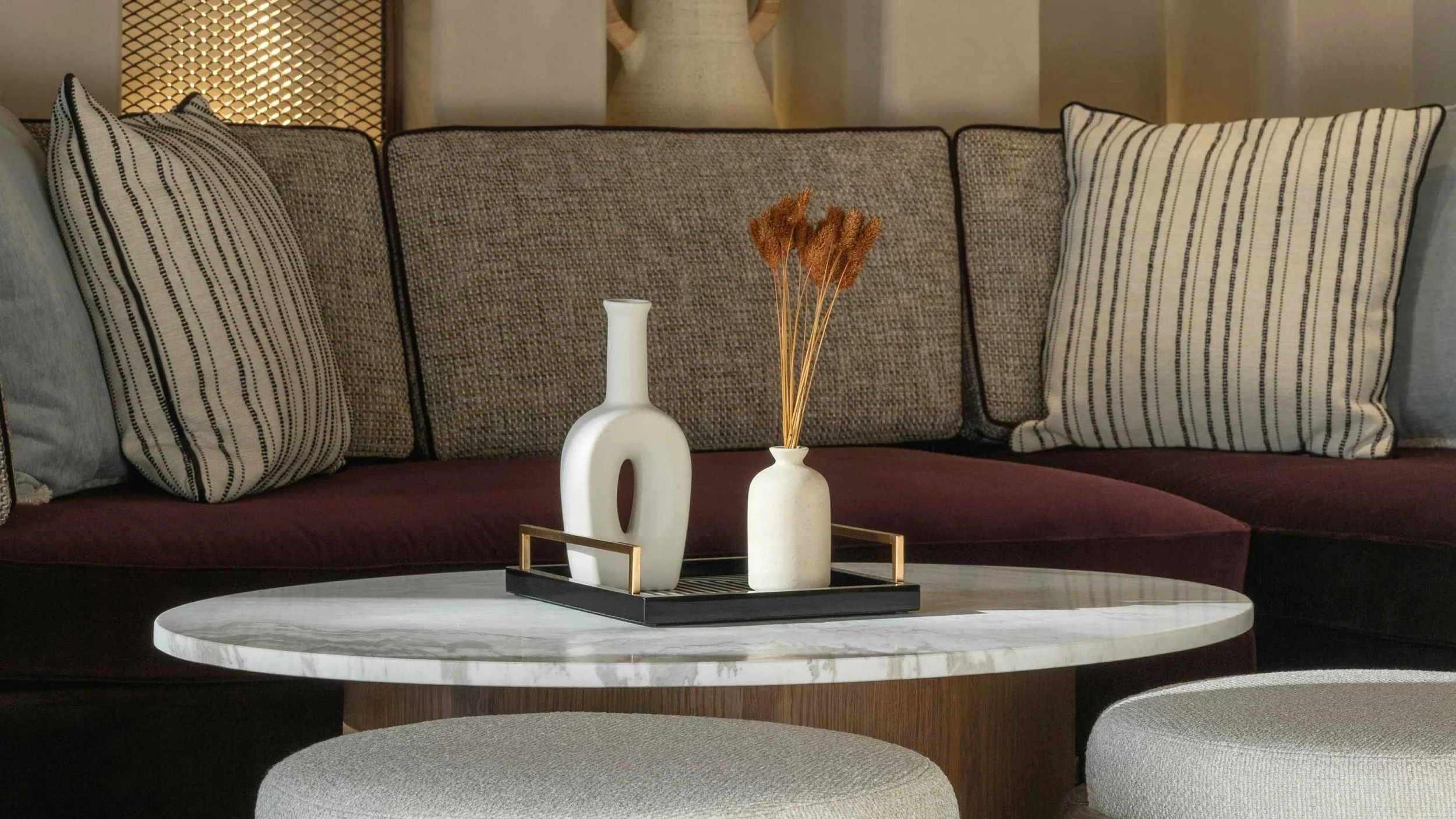 Living room with a purple sofa decorated with patterned pillows, a round marble coffee table holding two white vases with dried flowers, and a textured partition with a decorative sculpture in the background.