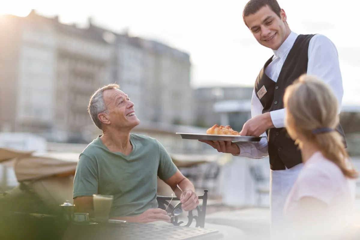 A man at an outdoor restaurant being served pizza by a waiter, with a smiling elderly man and a woman seated nearby.