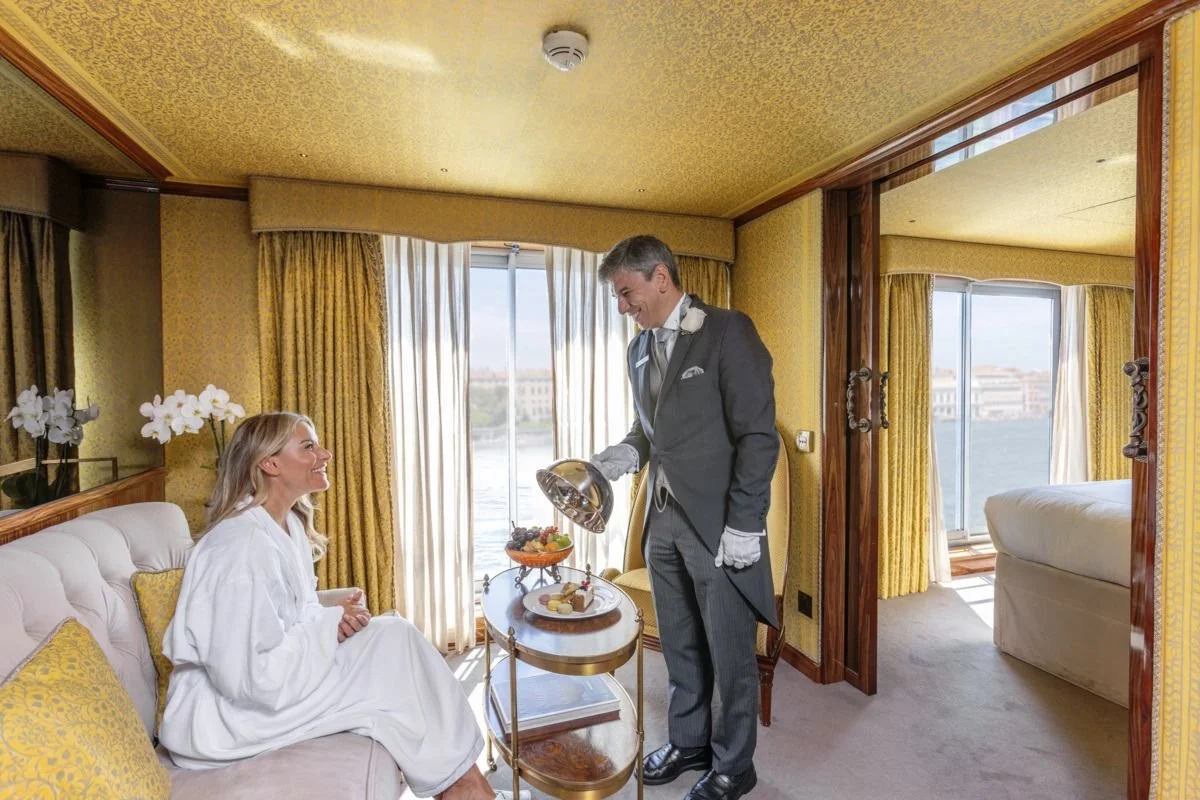 A woman sitting on a cream-colored sofa with yellow pillows, smiling at a man in a tuxedo. The man is serving breakfast from a silver tray. The room has yellow walls, large windows with yellow curtains, and a balcony with a view of the water and buildings.
