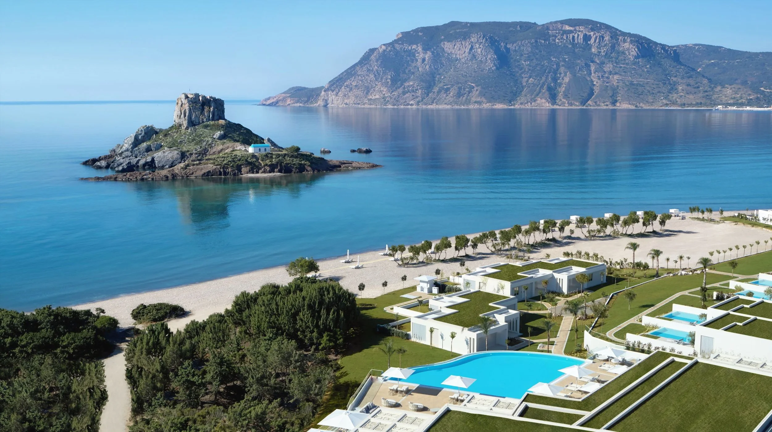 A coastal scene featuring a rocky island with a small white church and a bell tower, calm blue water, a sandy beach with umbrellas, green trees, modern white buildings with rooftop gardens and pools, and mountains in the background under a clear sky.