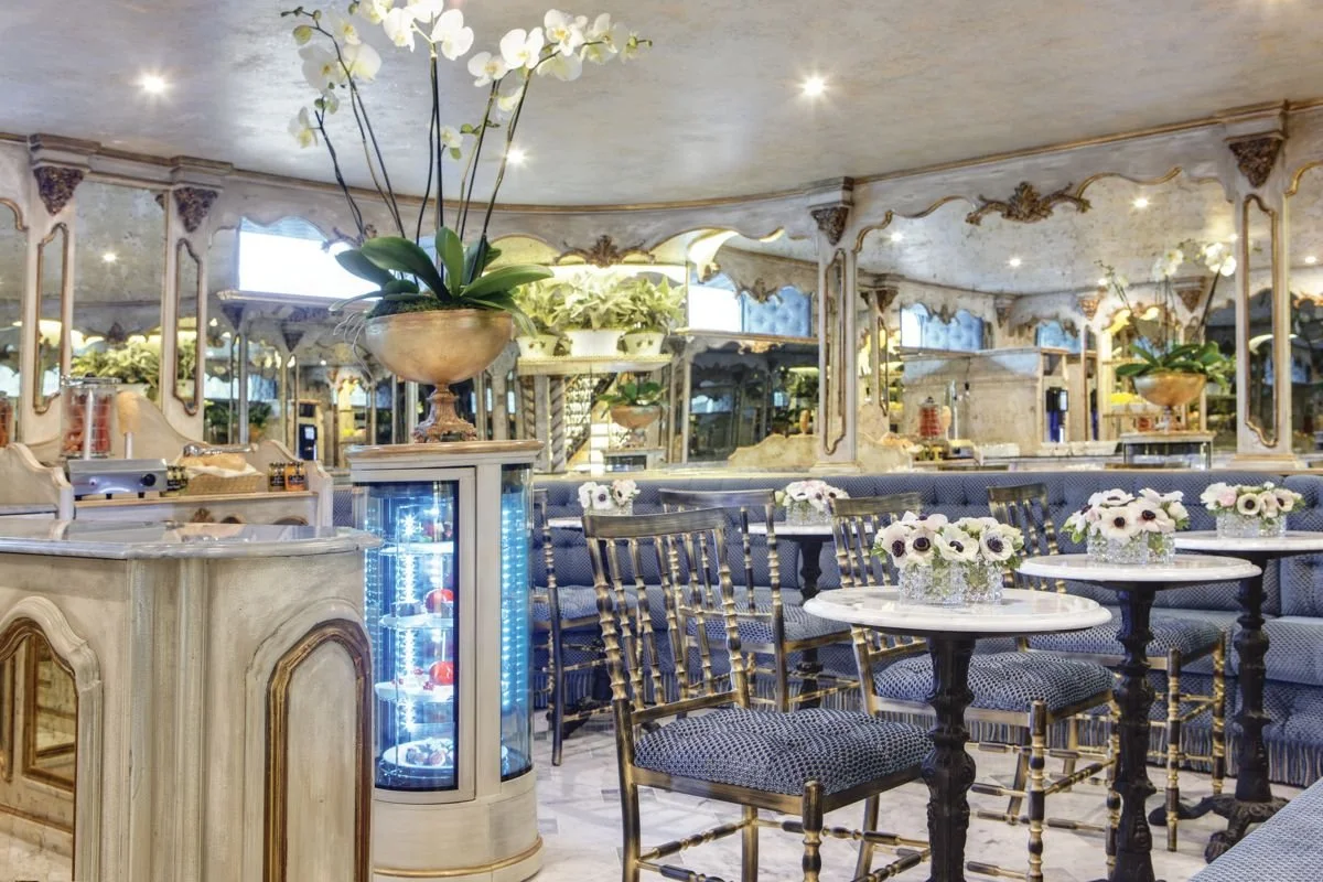 Elegant restaurant interior with marble tables, floral centerpieces, blue patterned chairs, mirrors, and decorative mirrors on the ceiling.