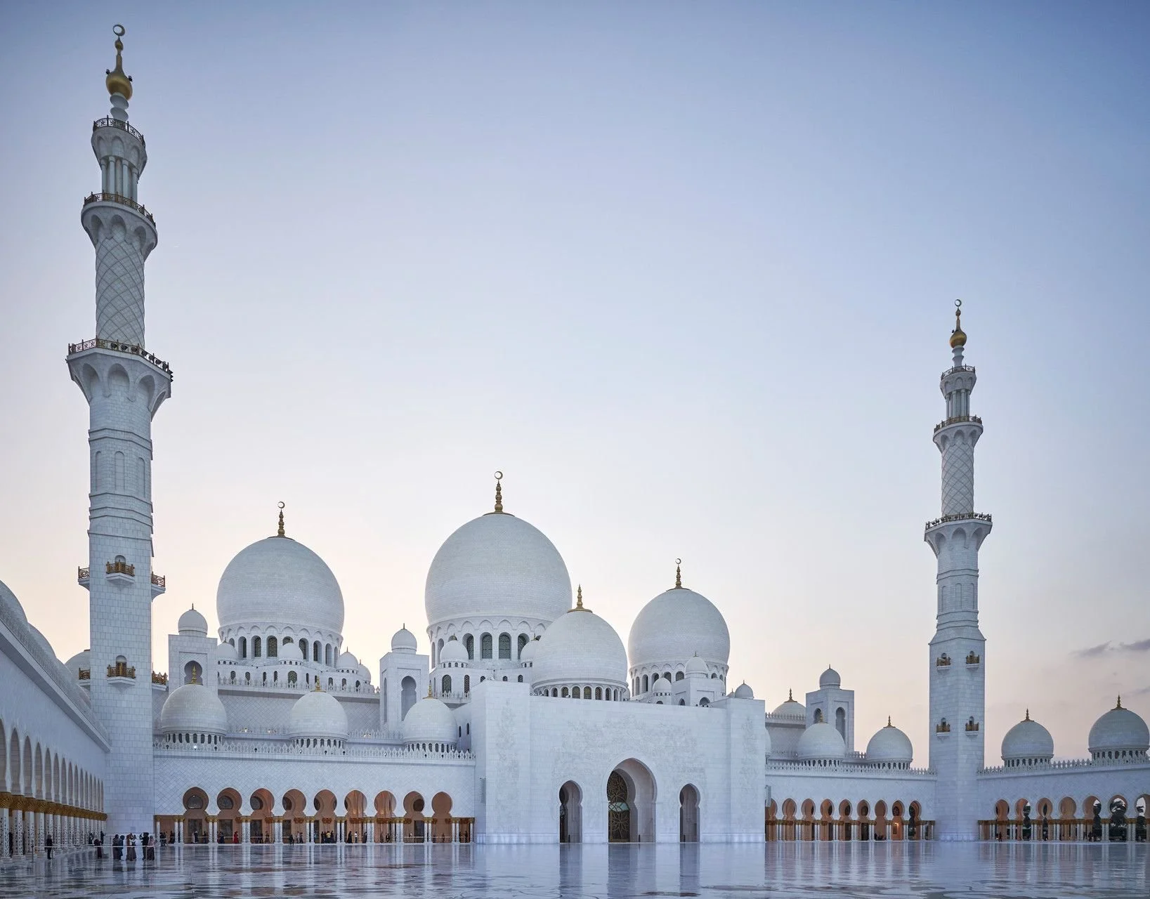 The image shows the Sheikh Zayed Grand Mosque in Abu Dhabi, United Arab Emirates, with white domes and minarets against a clear sky. The mosque's courtyard has a reflective marble floor with a few visitors visible.