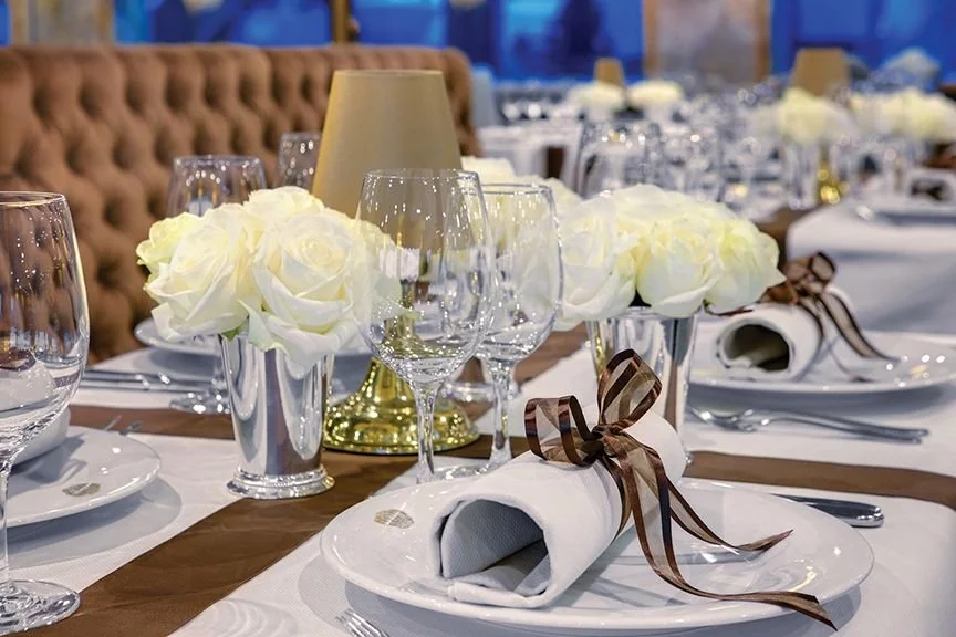 Elegant dining table setup with white roses in glass vases, wine glasses, white napkins tied with brown ribbons, and a lamp in the background.