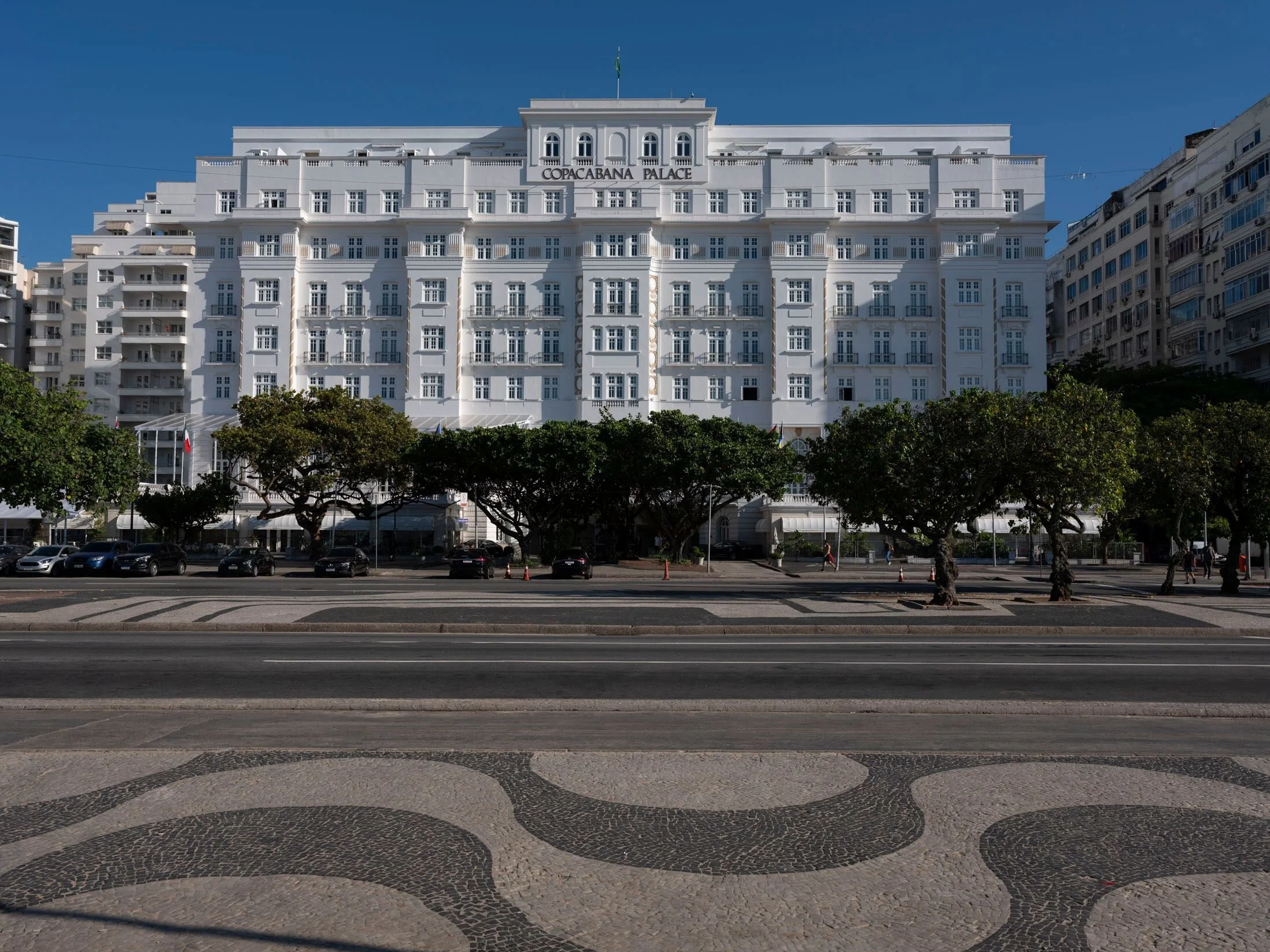 White historic hotel building with multiple balconies and the sign 'Copacabana Palace' on the front, situated in an urban area with neighboring buildings on either side, trees, and parked cars in front.