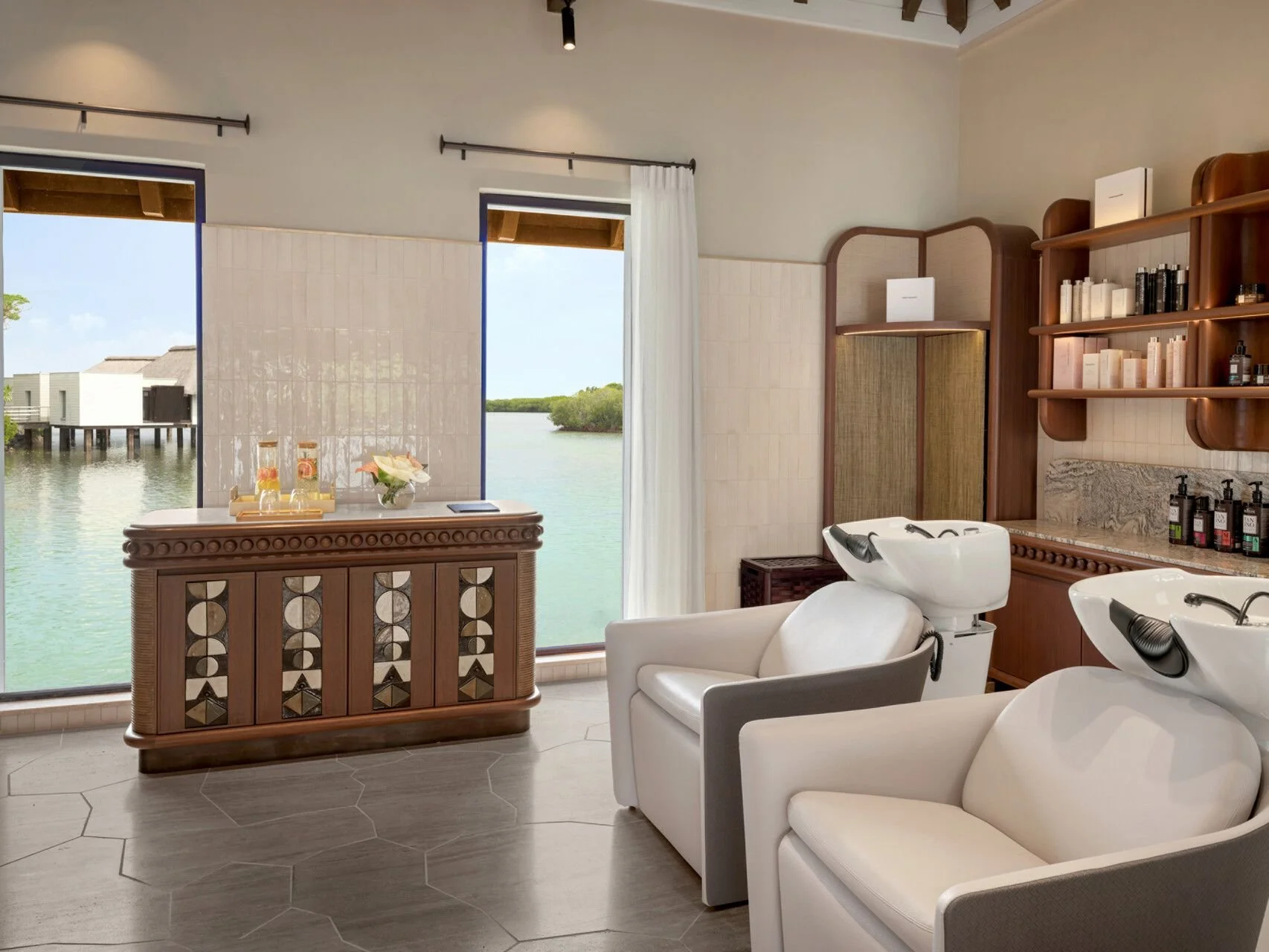 A modern hair salon with two white sinks and chairs, wooden shelves with hair products, and large windows overlooking water and overwater bungalows in the background.