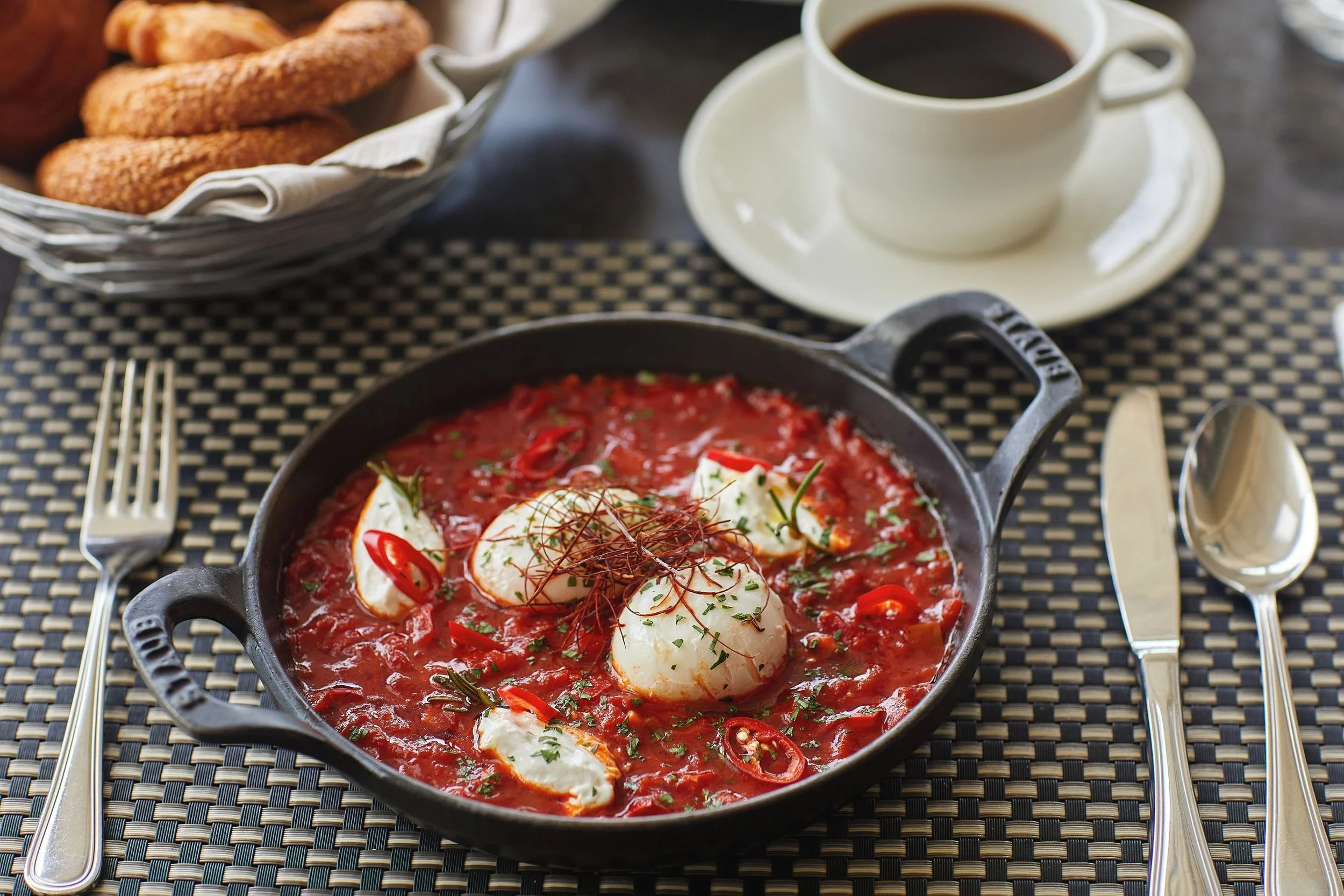 A skillet of eggs benedict with red chili slices, herbs, and a tomato-based sauce. In the background, there is a bowl of toast or bread, a cup of black coffee on a saucer, and silverware on a dark woven placemat.