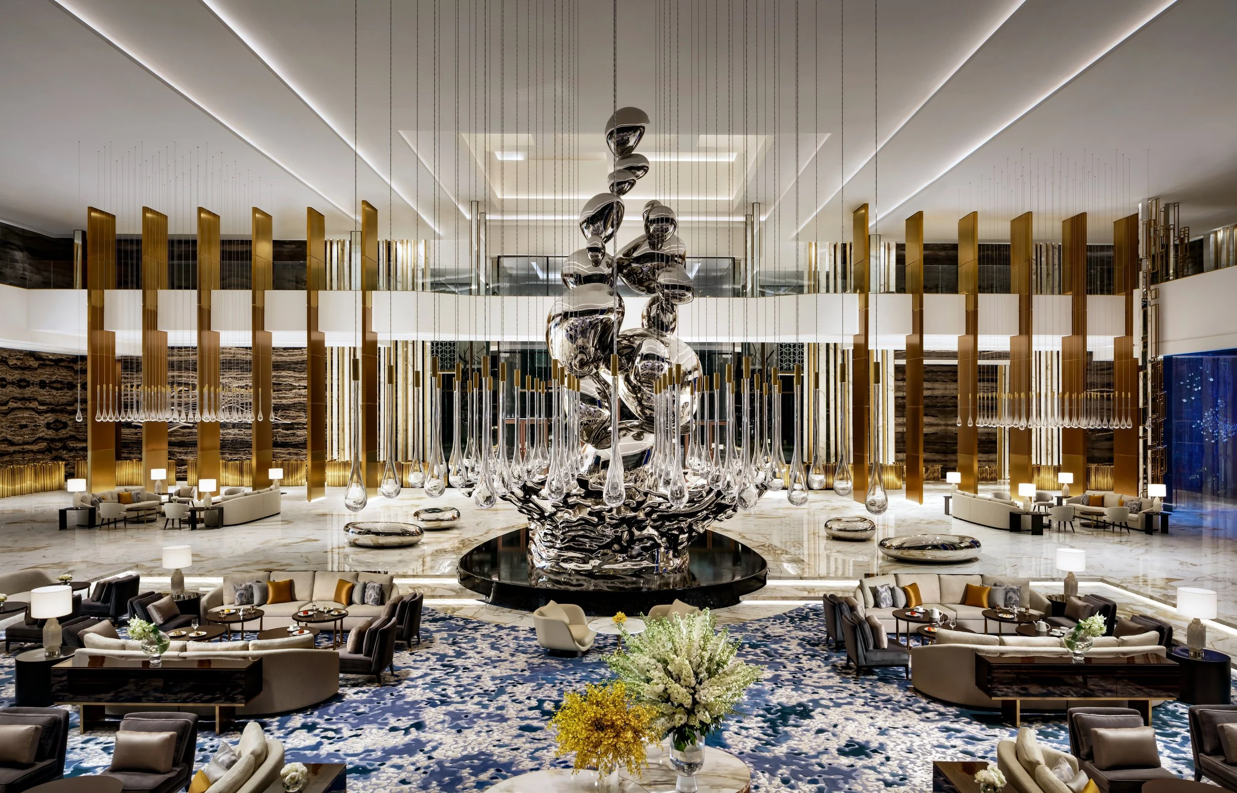 Elegant hotel lobby with high ceiling, modern silver and black abstract sculpture, gold and white vertical decorative panels, marble floor, seating areas with sofas and chairs, and floral arrangements.