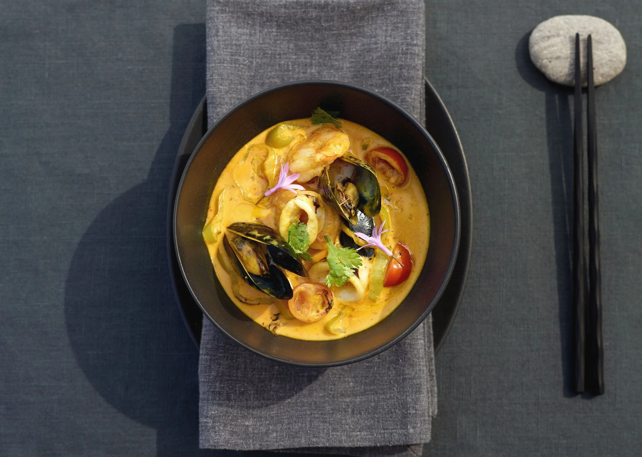 A bowl of seafood curry with shrimp and mussels garnished with cilantro and edible flowers, served on a dark textured table with chopsticks on a stone and fabric placemat.