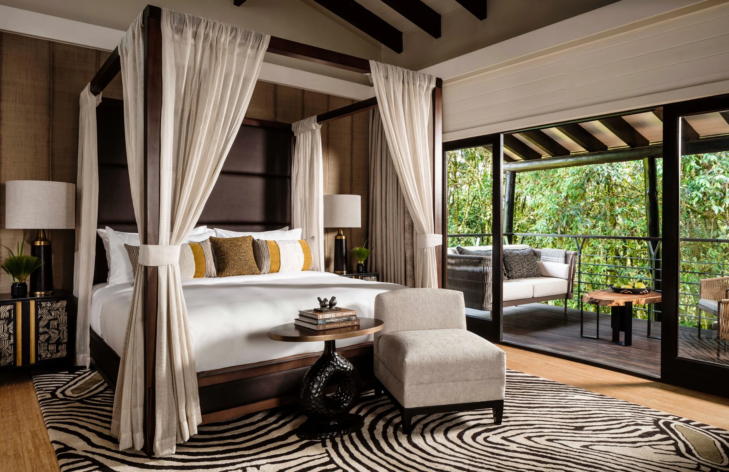 A luxurious bedroom with a four-poster bed, beige curtains, and pillows, next to an outdoor balcony with green foliage, a seating area, and a small table.