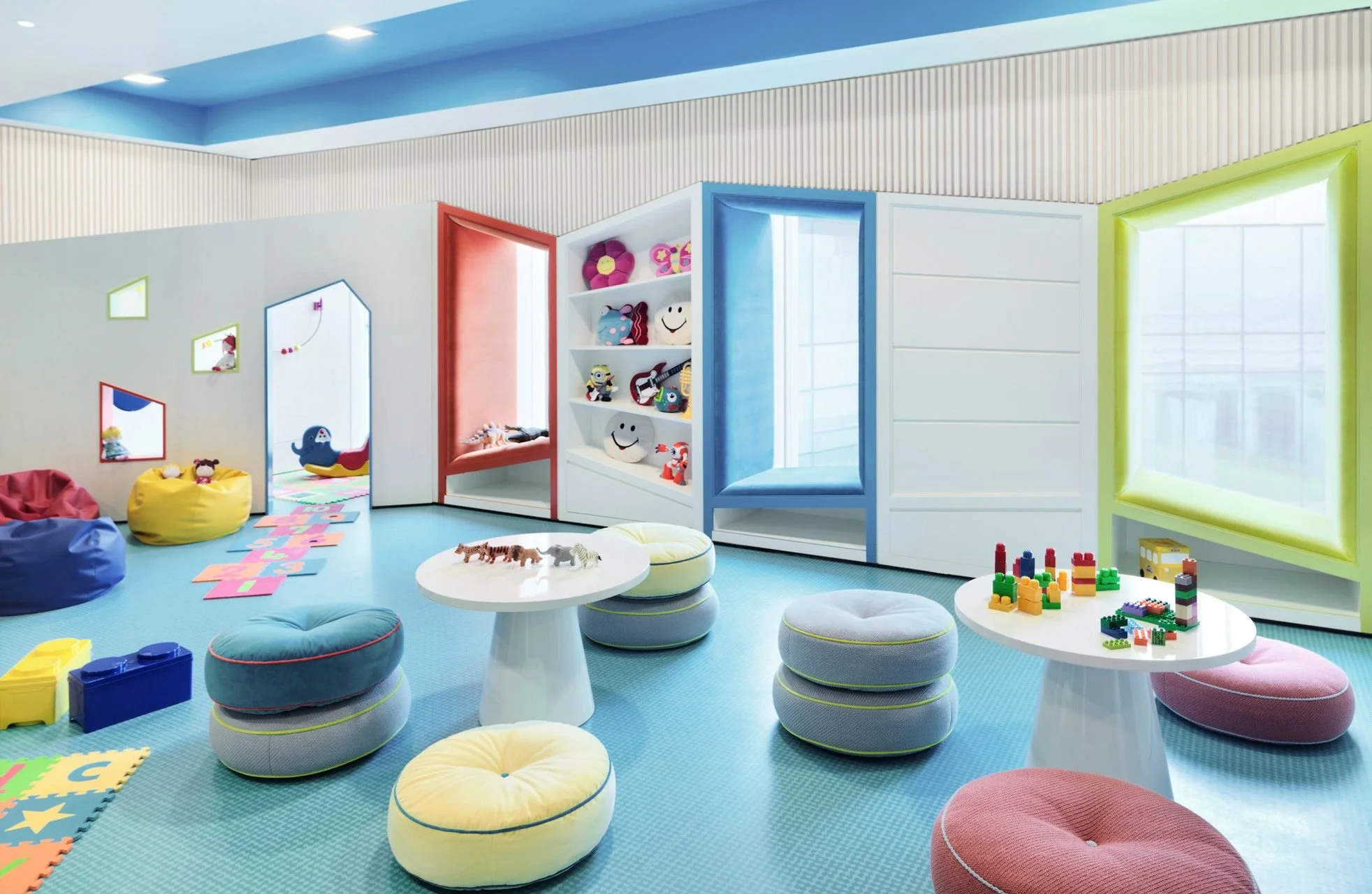 Colorful children's playroom with bean bag chairs, round cushions, and tables with toys and building blocks. Bright windows and decorated walls create a cheerful atmosphere.