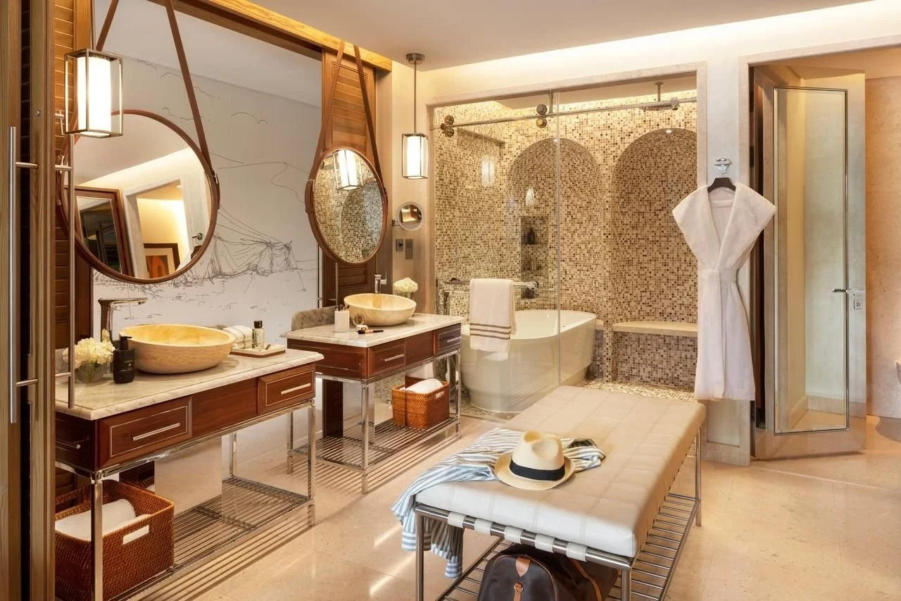 Luxurious hotel bathroom with two vessel sinks on a marble countertop, round mirrors above each sink, and modern lighting fixtures. In the background, there is a glass-enclosed shower area with mosaic tile walls, a separate bathtub, and a white bathrobe hanging on a hook. A bench with a hat and striped clothing, along with a black bag, is in the foreground.