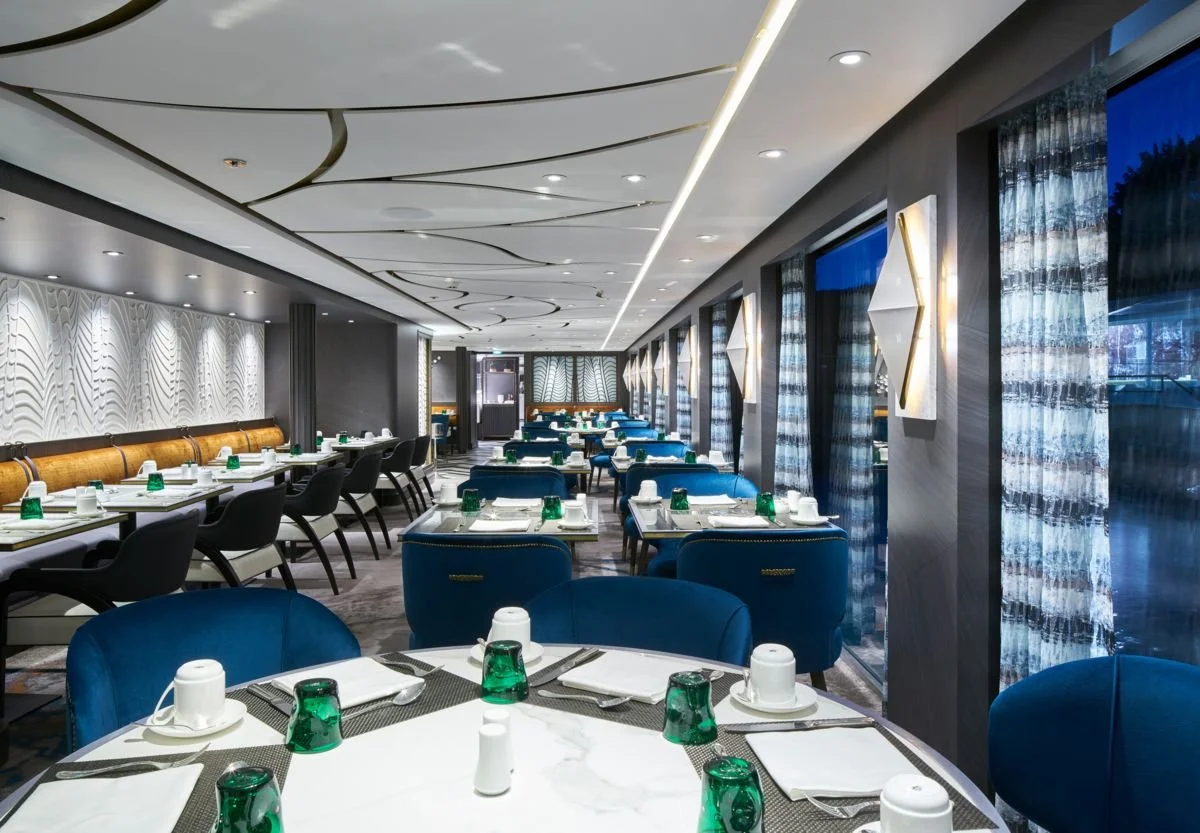 Modern restaurant interior with blue chairs, white marble tables set with cups, plates, glasses, and silverware, large windows with patterned curtains, and decorative wall lighting.