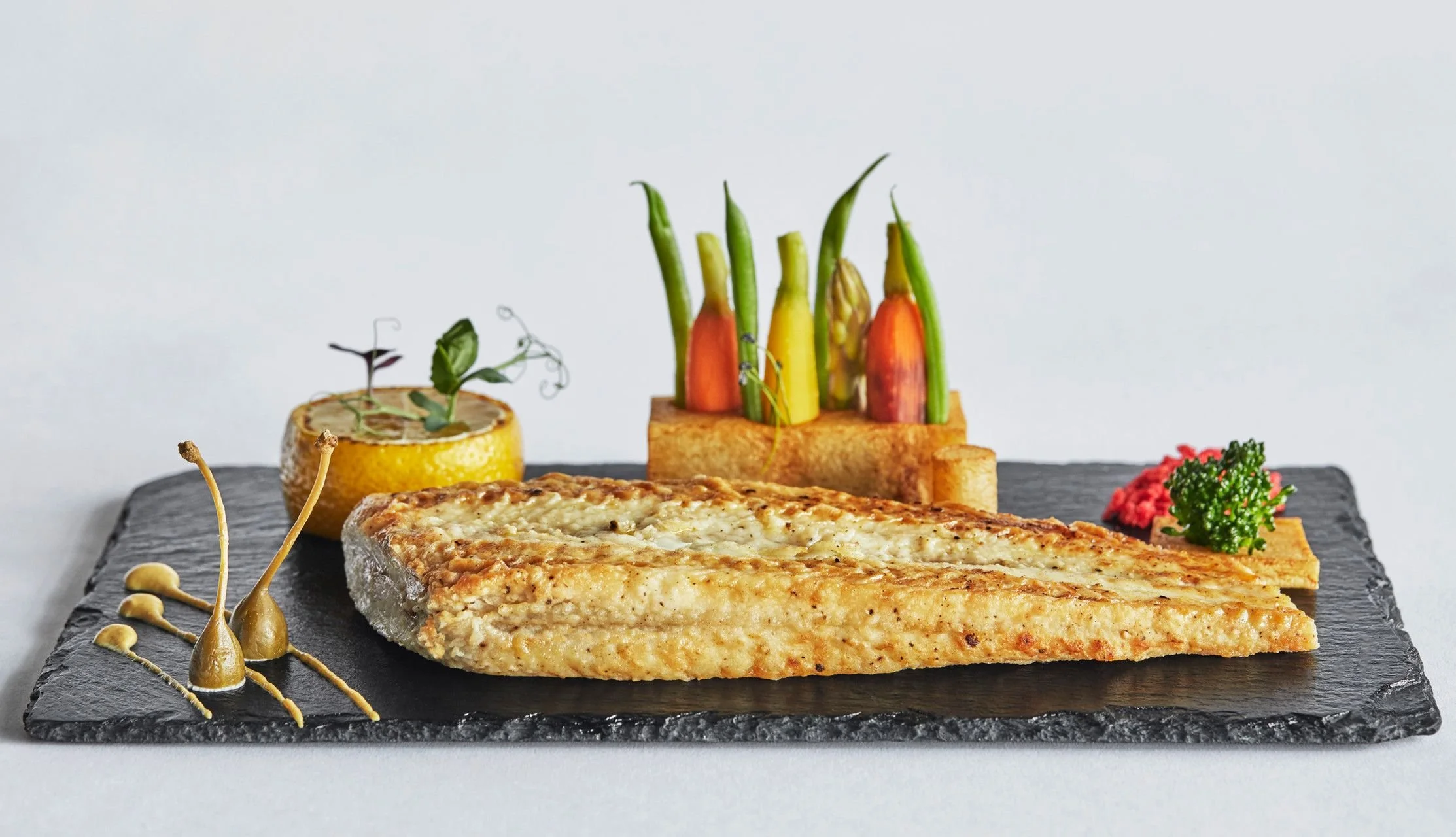 A slice of baked fish with a golden crust served on a black slate platter, garnished with microgreens, and accompanied by roasted vegetables including carrots, zucchini, and a half lemon with a decorative sprig, on a light background.