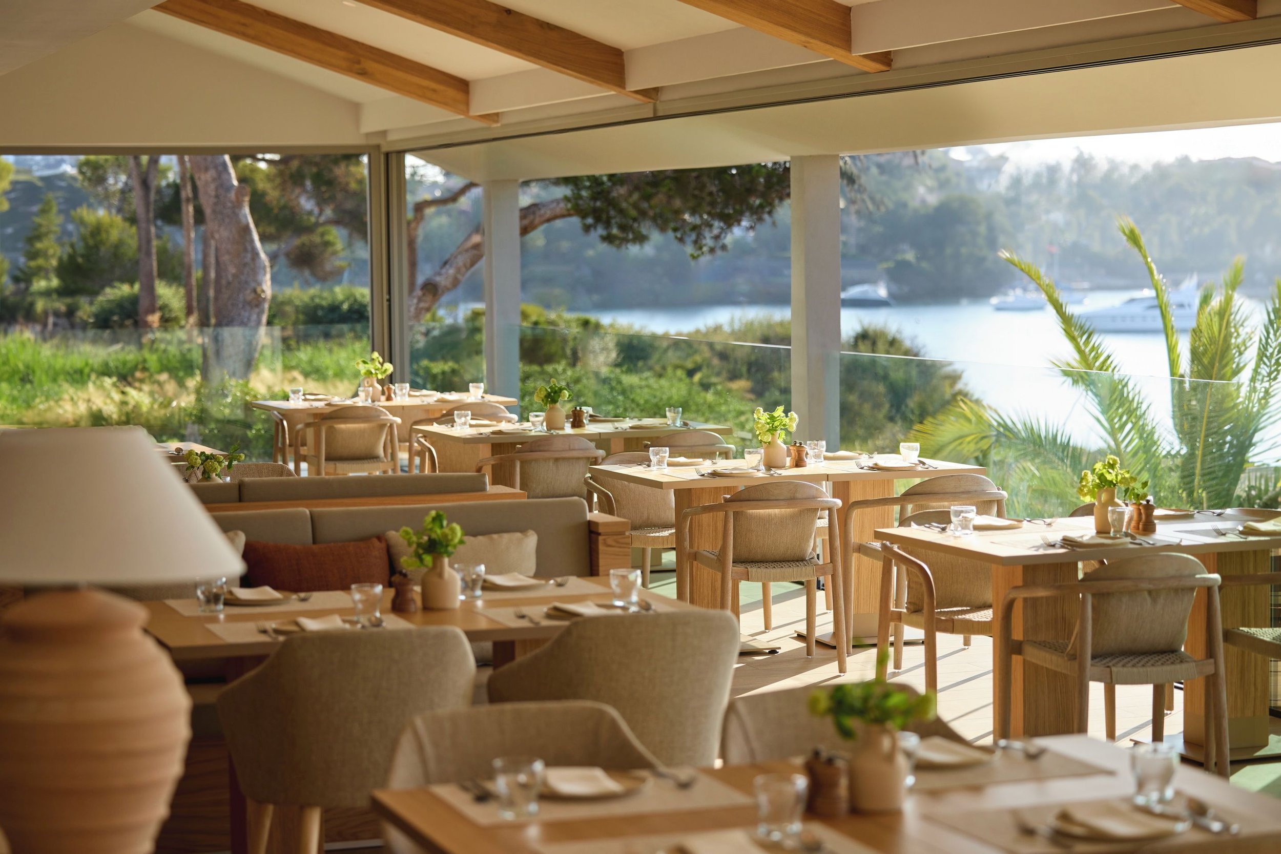 A bright, modern restaurant with large windows overlooking trees and water, wooden beams on the ceiling, and tables set with plates, glasses, and small vases of green flowers.