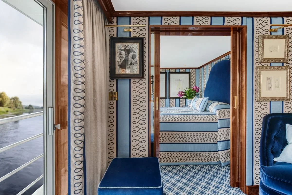 Interior of a boat or luxury cabin with patterned blue and gold wallpaper, a doorway leading to a bedroom with a bed and a large blue headboard, framed artwork on the walls, a window with beige curtains, and a view of water outside.