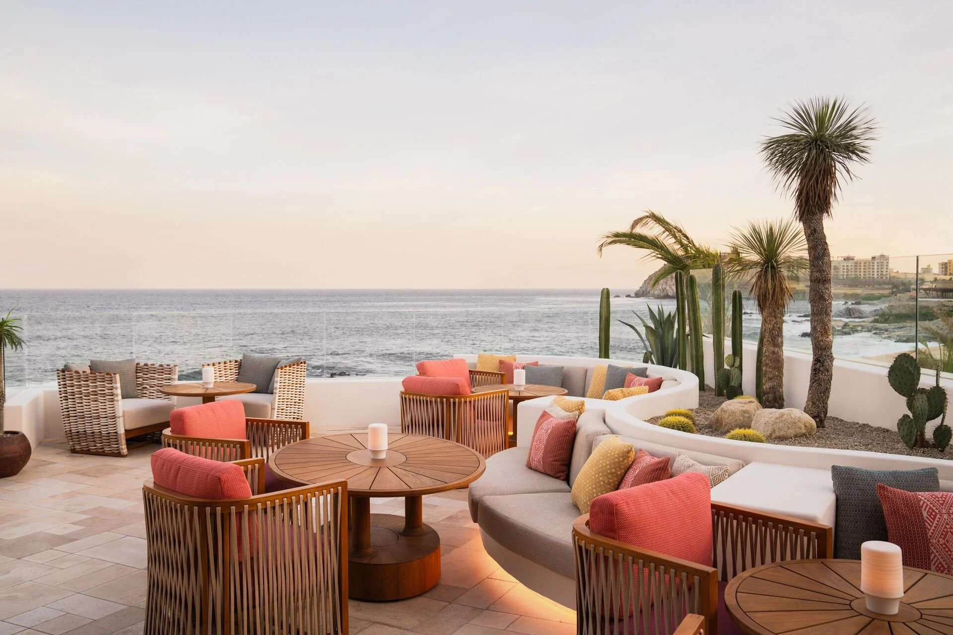 Outdoor patio with wicker chairs, cushioned seating, and wooden tables overlooking the ocean. Palm trees and cacti decorate the area, with a view of cliffs and a coastal town in the distance during sunset.