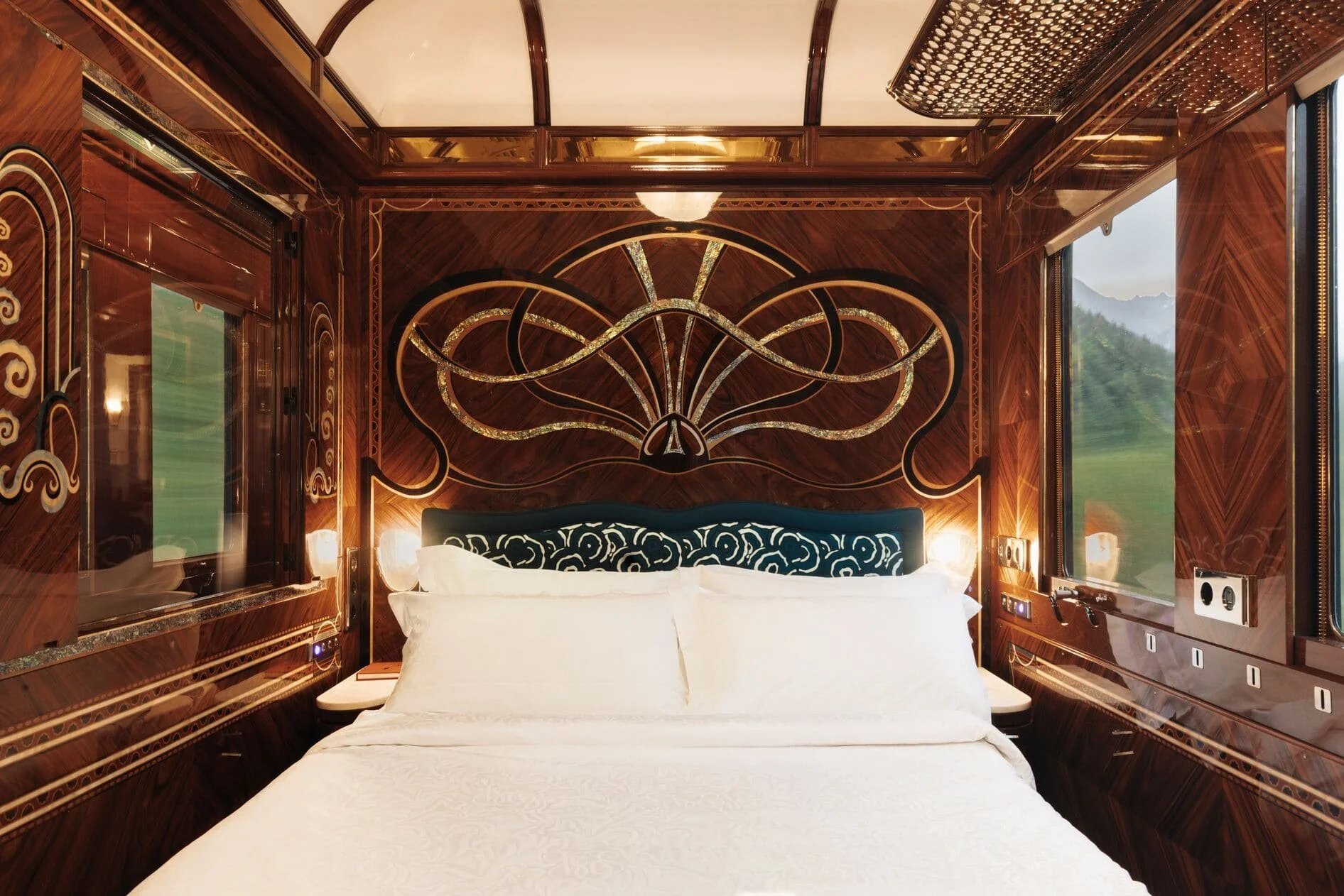 Interior of a luxurious bedroom with dark polished wood walls and headboard, featuring intricate gold detail and a large window showing mountains and trees outside.