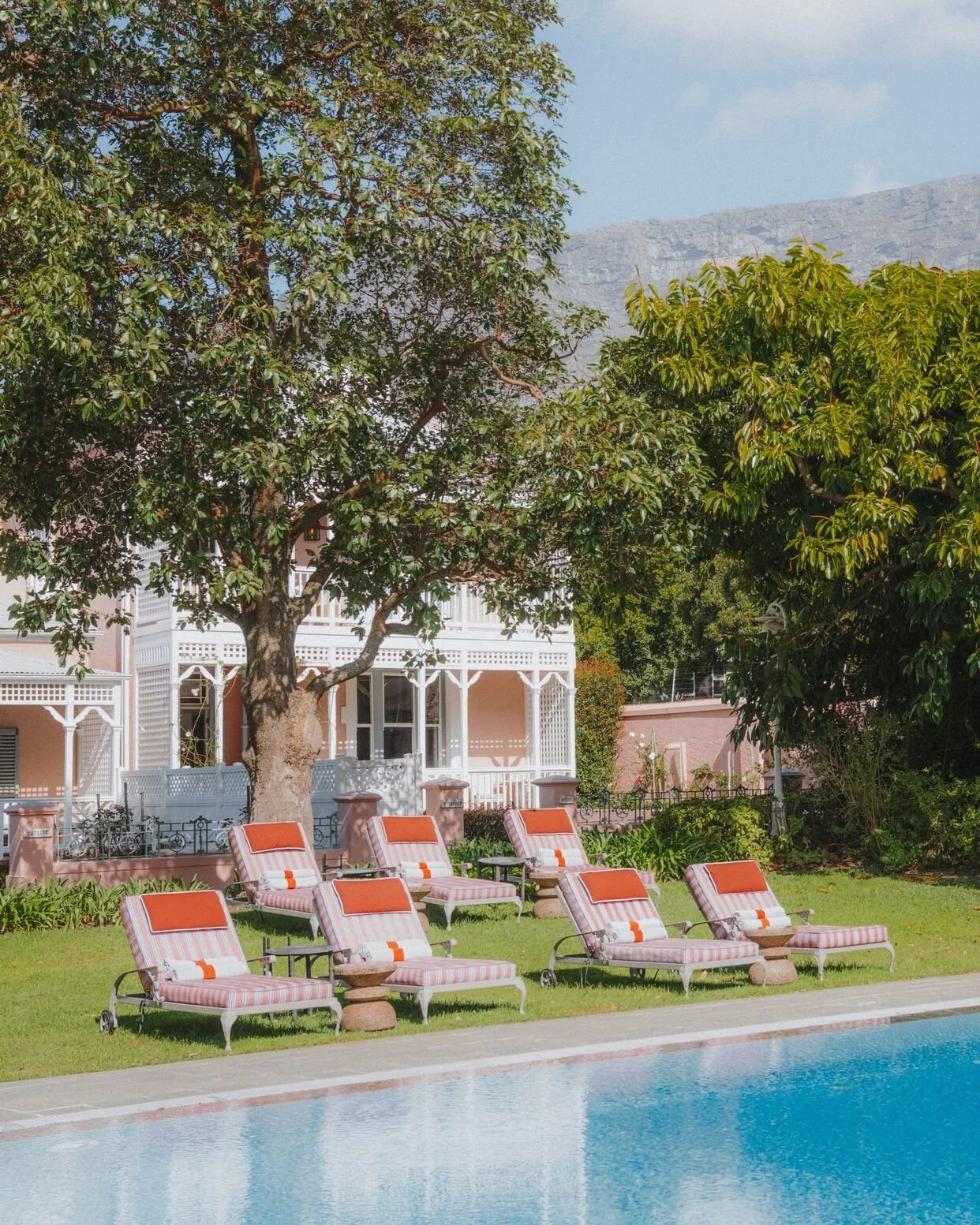 Poolside scene with multiple pink-striped lounge chairs with orange cushions, green lawn, large trees, a pink house with white decor, and a mountain in the background.