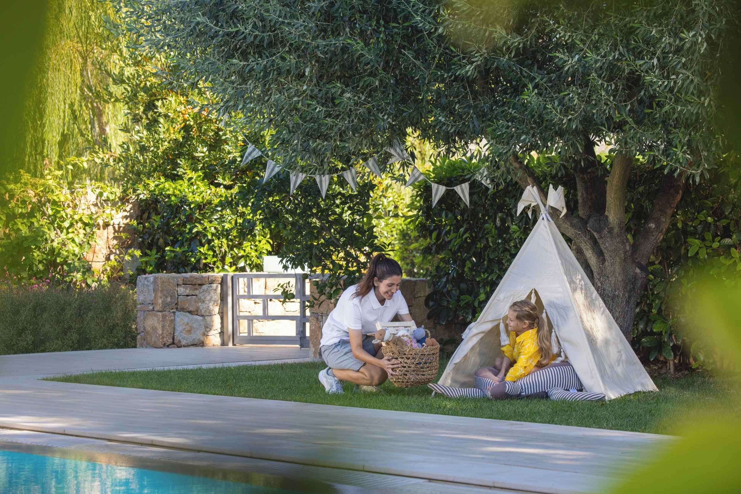 A woman and a young girl are sitting outside near a small white tent under a large tree. The woman is smiling while holding a basket of toys, and the girl is sitting on a blanket, dressed in a yellow raincoat, looking at the woman. The scene is set in a lush, green garden with sunlight filtering through the leaves.