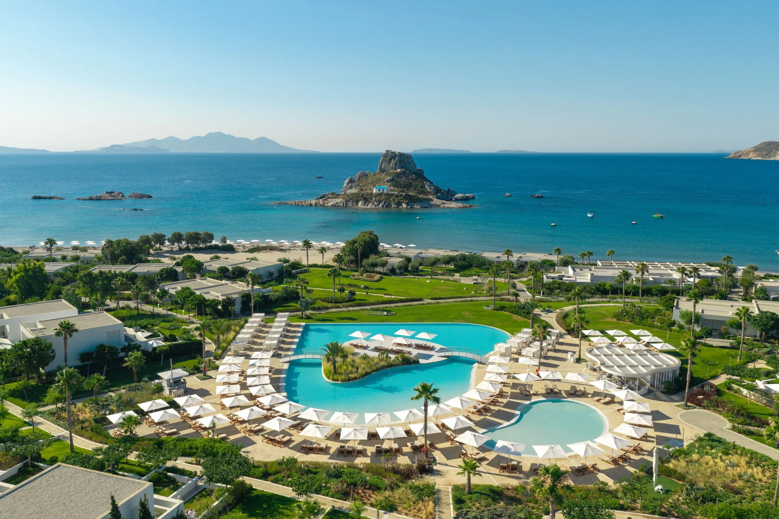 Aerial view of a resort with a large swimming pool and lounge chairs, surrounded by palm trees, overlooking the ocean with small islands and mountains in the background.