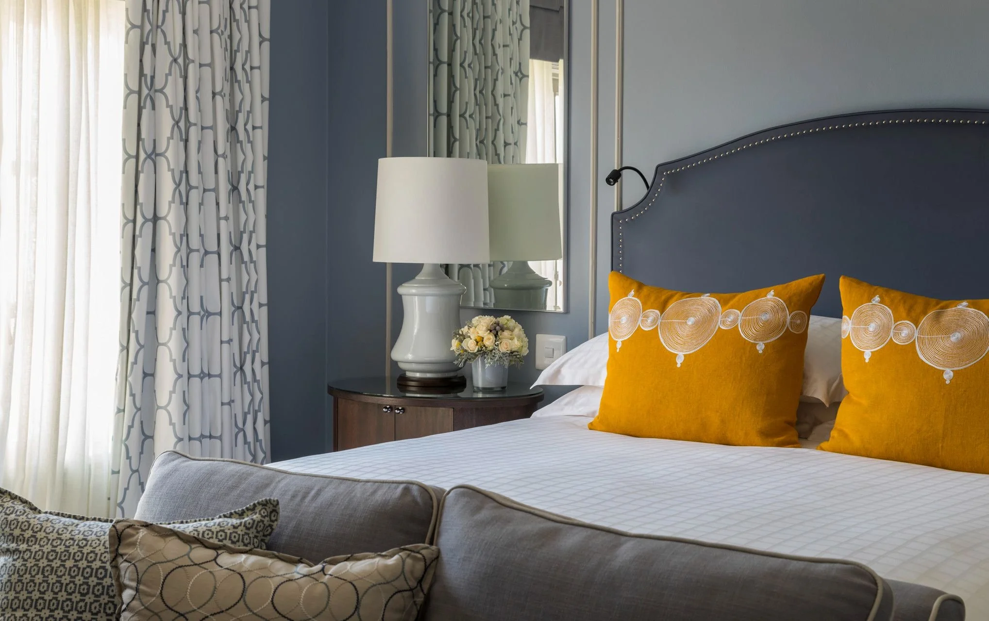 A bedroom with a bed, yellow pillows with circular patterns, a side table with a flower arrangement, a lamp, a mirror, and curtains with a geometric pattern.