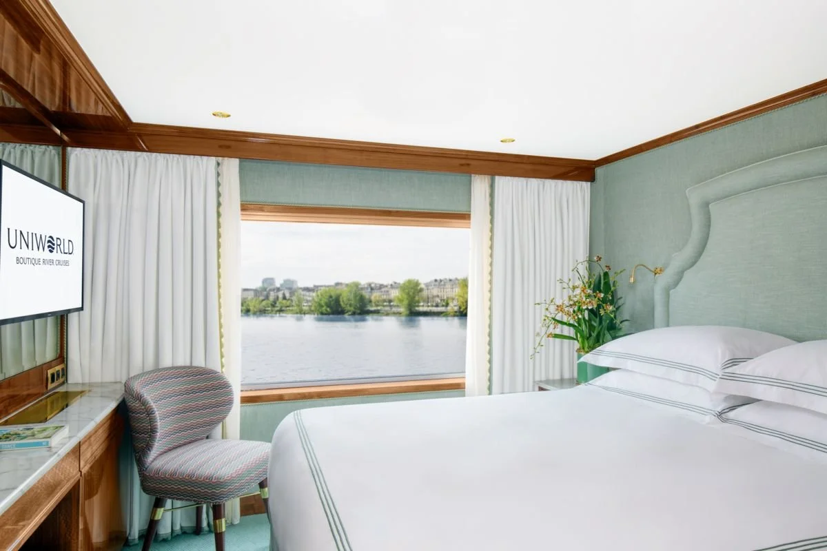 Hotel room with a bed, a window viewing a river and cityscape, a TV, a patterned chair, and a vase with flowers