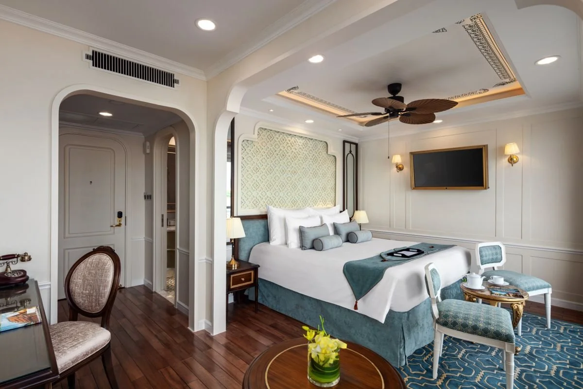 A luxurious hotel room with a large bed, side tables with lamps, a wall-mounted TV, a ceiling fan, and seating area with chairs and a small table, featuring elegant decor and hardwood floors.