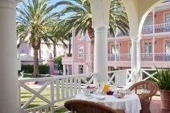 Outdoor dining table set with plates, glasses, and a white tablecloth on a patio with white columns, pink and purple buildings, and palm trees in the background.