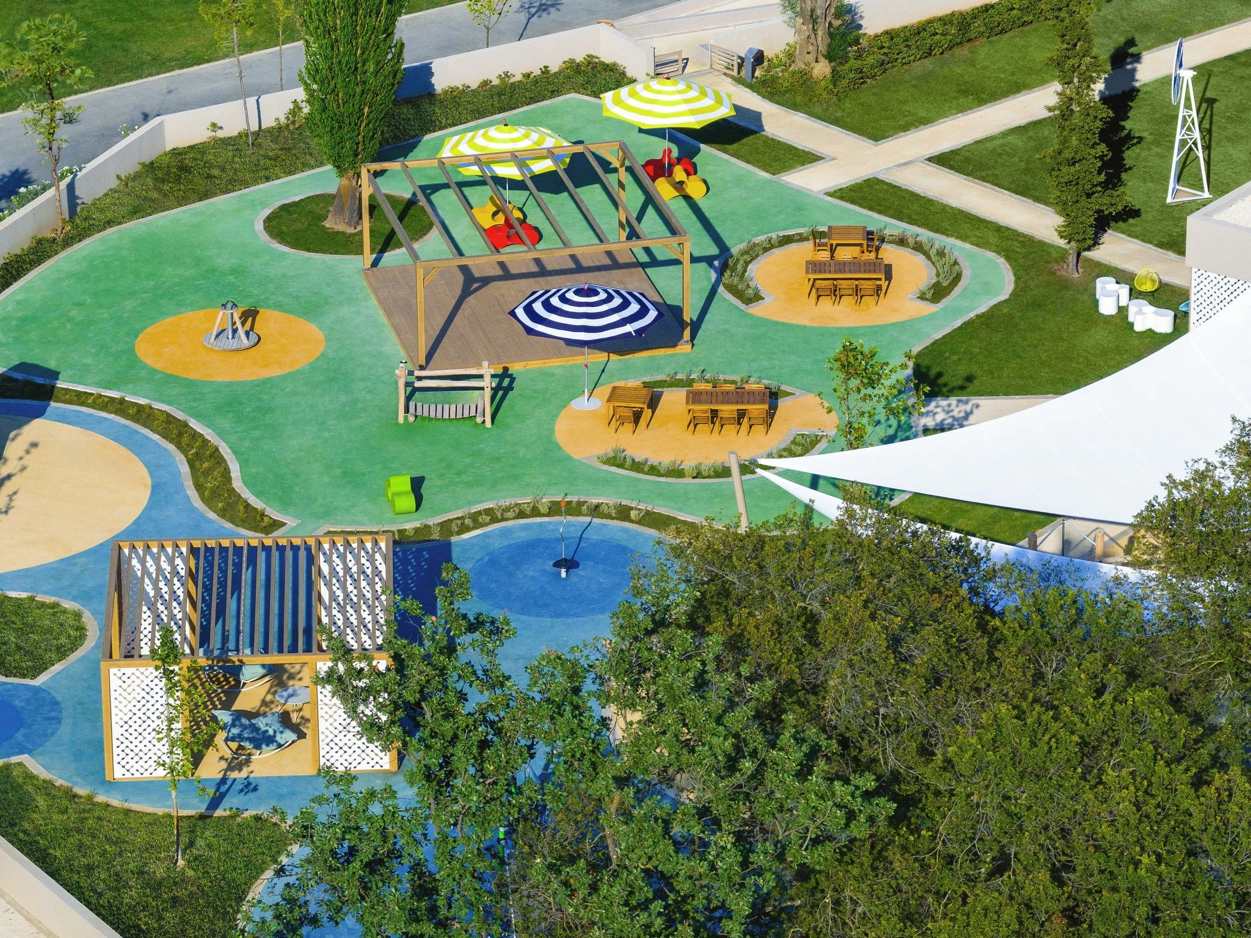 A playground with various equipment including swings, a sandbox, and shaded areas, surrounded by grass and trees.