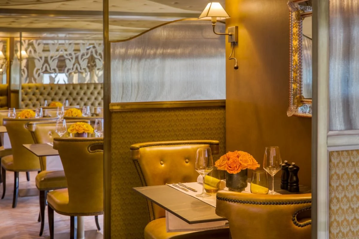 An elegant restaurant interior with yellowish upholstered chairs, a table set with wine glasses, yellow flowers in a pot, and a mirror on the wall.