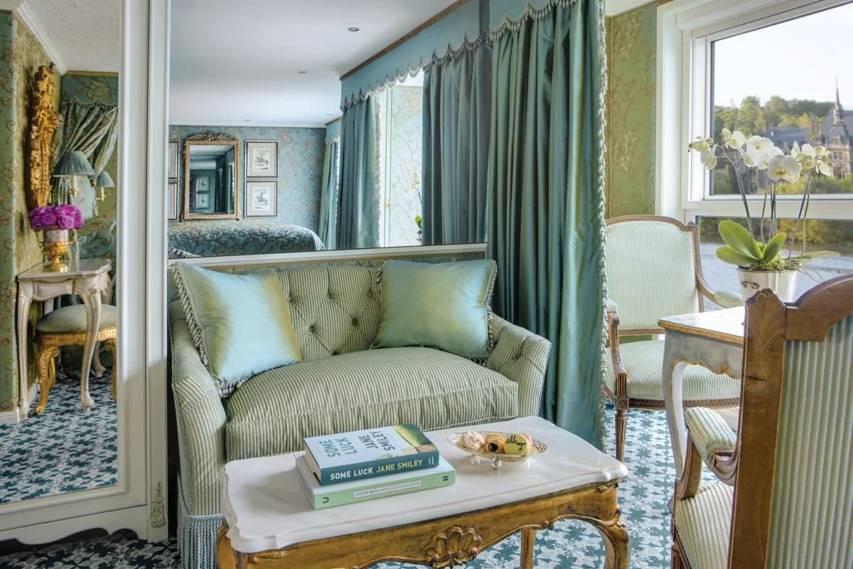 A brightly colored living room decorated in pastel green and gold tones with elegant vintage furniture, including a sofa, armchairs, and a mirror. There are books and decorative items on the coffee table, and sunlight coming through the windows.