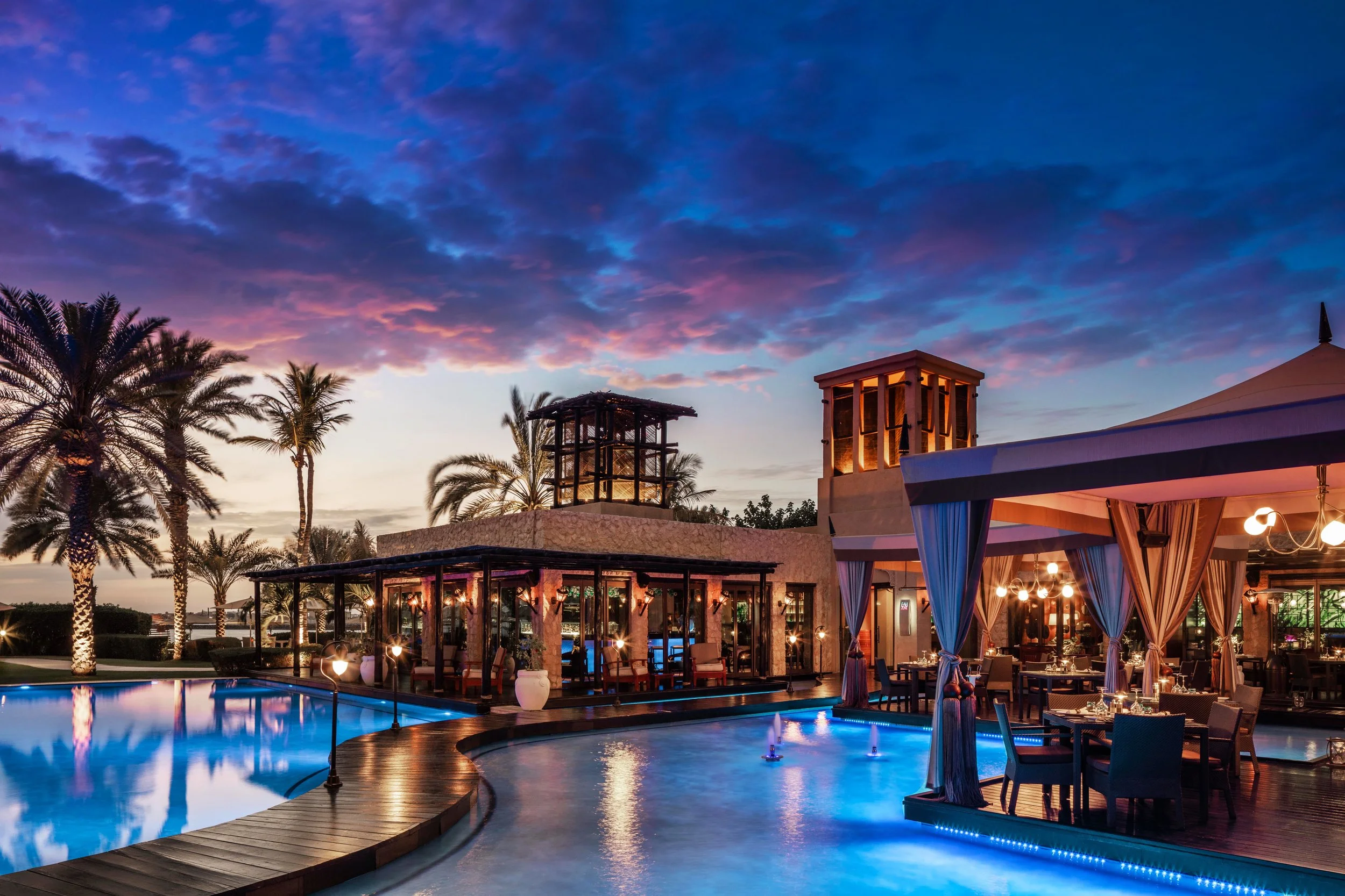 A scenic view of a swimming pool area during sunset with palm trees, outdoor seating, and a building with lit windows and a rooftop structure under a colorful sky with purple and orange clouds.
