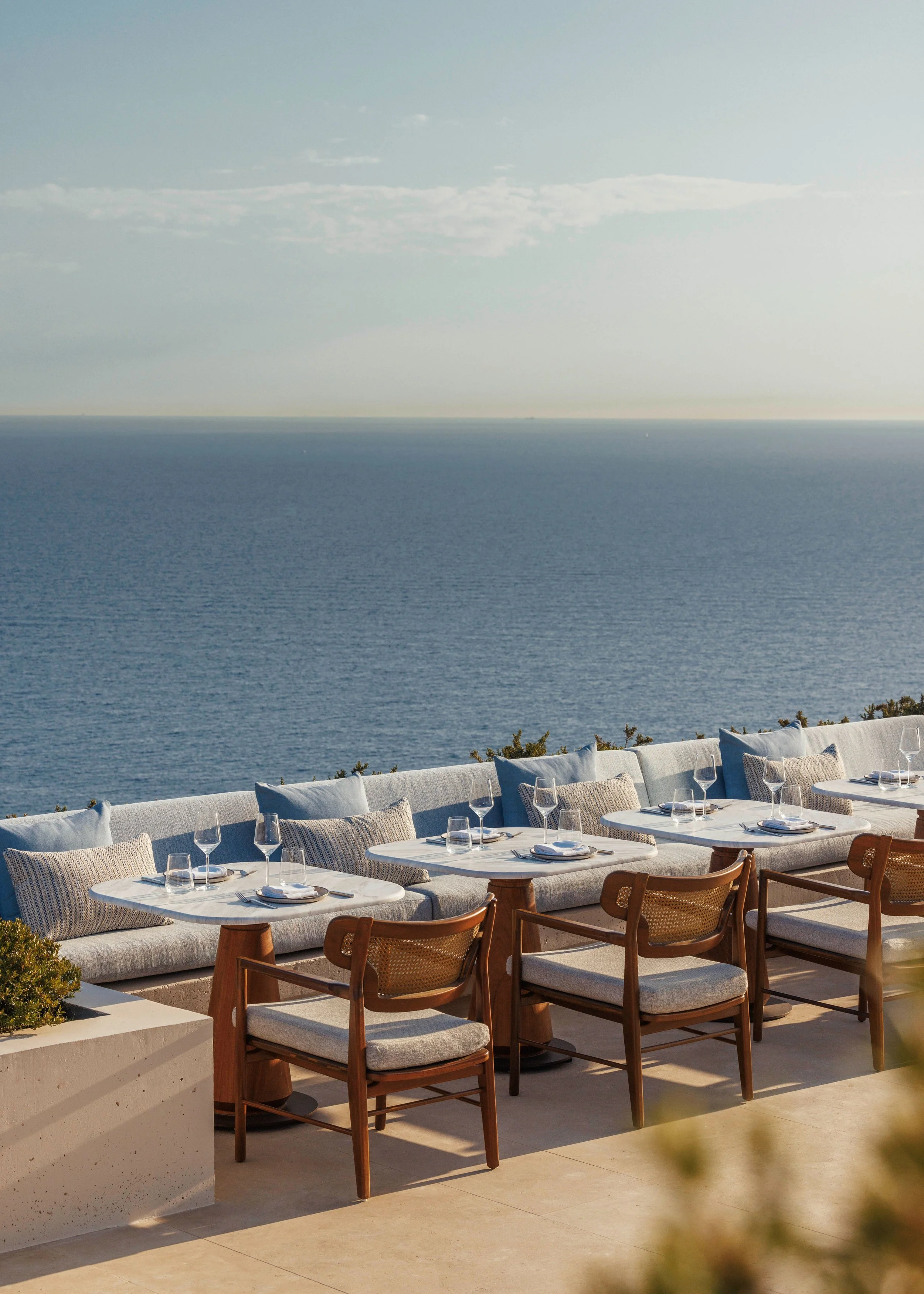 Outdoor dining area with white cushioned seating and wooden chairs, set with plates, glasses, and napkins, overlooking the ocean with a pale blue sky and some clouds.