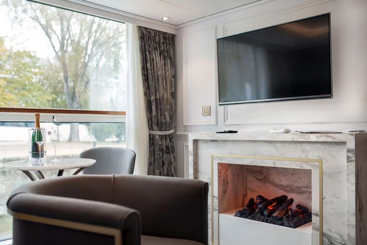 A cozy living room scene with a marble fireplace, a mounted flat-screen TV, a small round table with a bottle of champagne and two glasses, a gray upholstered armchair, and large windows showing trees outside.