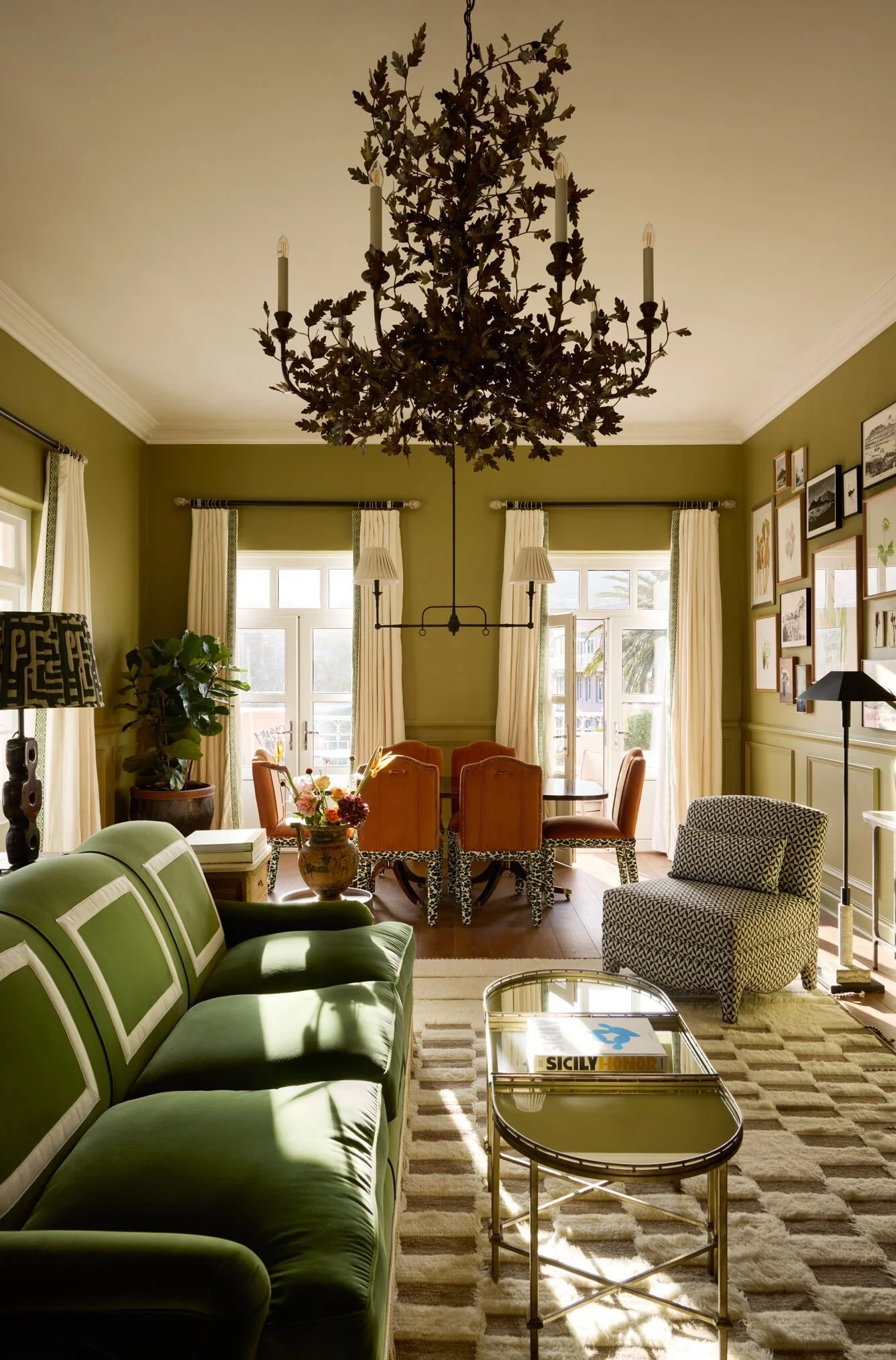Living room with green sofa, side tables, a patterned armchair, a coffee table with magazines, and a chandelier hanging from the ceiling. Large windows let in natural light, and walls are decorated with framed artwork.