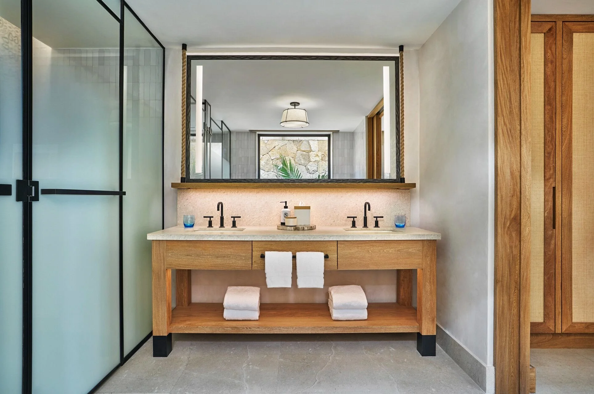 Modern bathroom with dual sinks, large mirror, glass shower enclosure, wooden accents, and minimal decor.