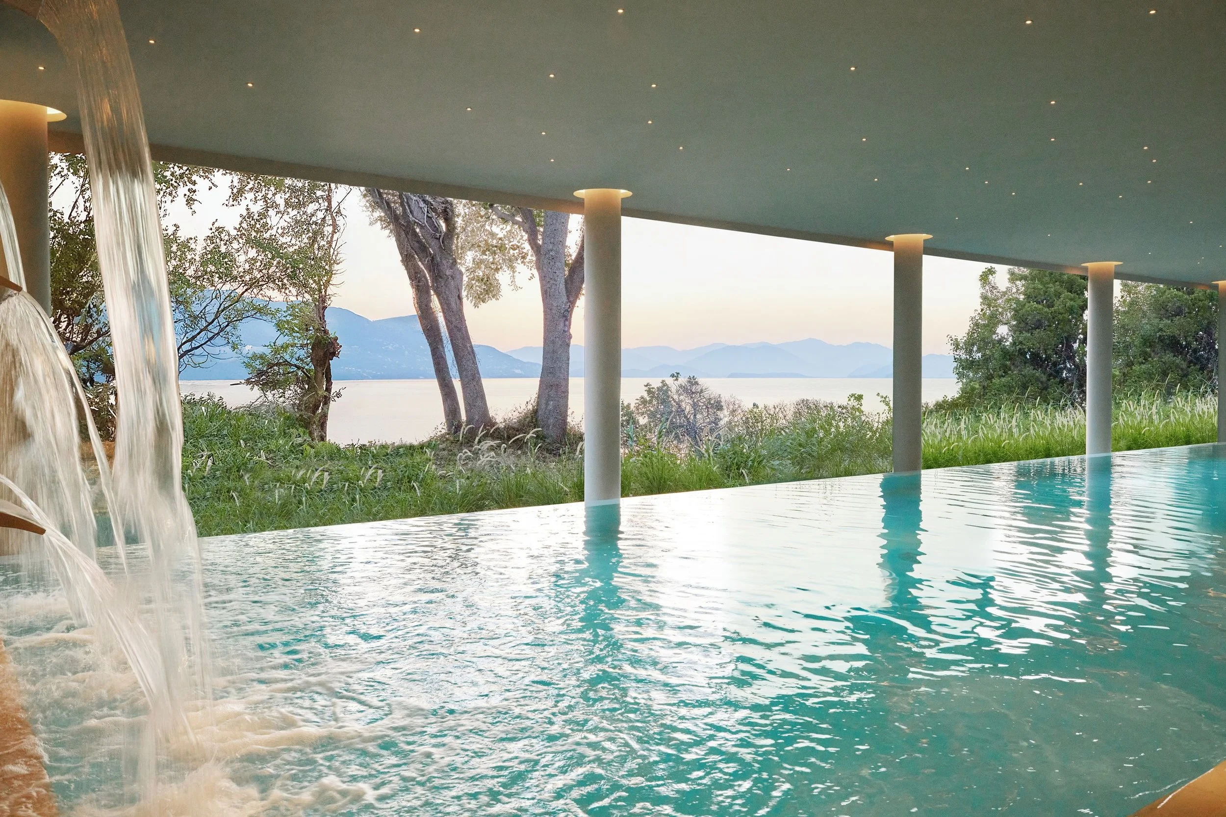 Indoor swimming pool with water fountain feature and floor-to-ceiling windows showing trees, mountains, and a lake outside.